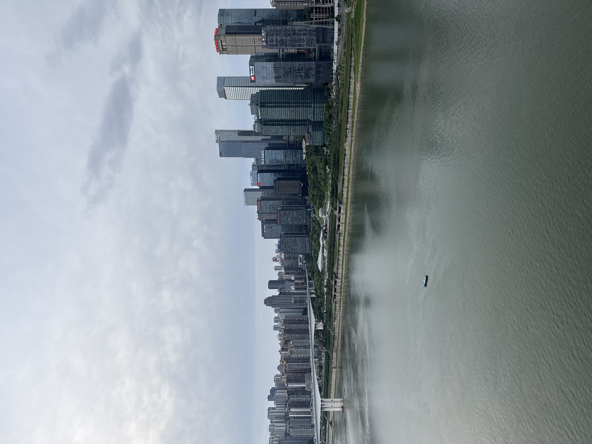 The image depicts a cityscape in Yuzhong District, China, with tall buildings along a riverbank. The river is gray-green in color and has a boat on it. A bridge spans the river in the foreground. The sky is overcast.