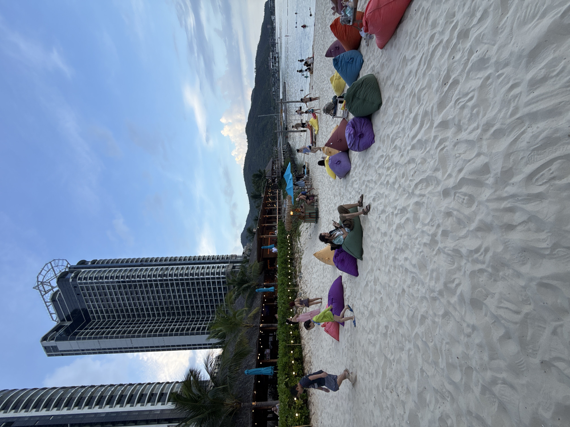 The scene is a beach in Bac Nha Trang Ward, Vietnam. In the background, there is a mountain and the ocean with some people swimming. A tall modern high-rise building stands on the left side of the image. A beachfront establishment, likely a restaurant or bar, runs along the sand. On the sand there are colorful beanbag chairs, with people relaxing and interacting on them. A child is walking towards the camera. The sky is partially cloudy.