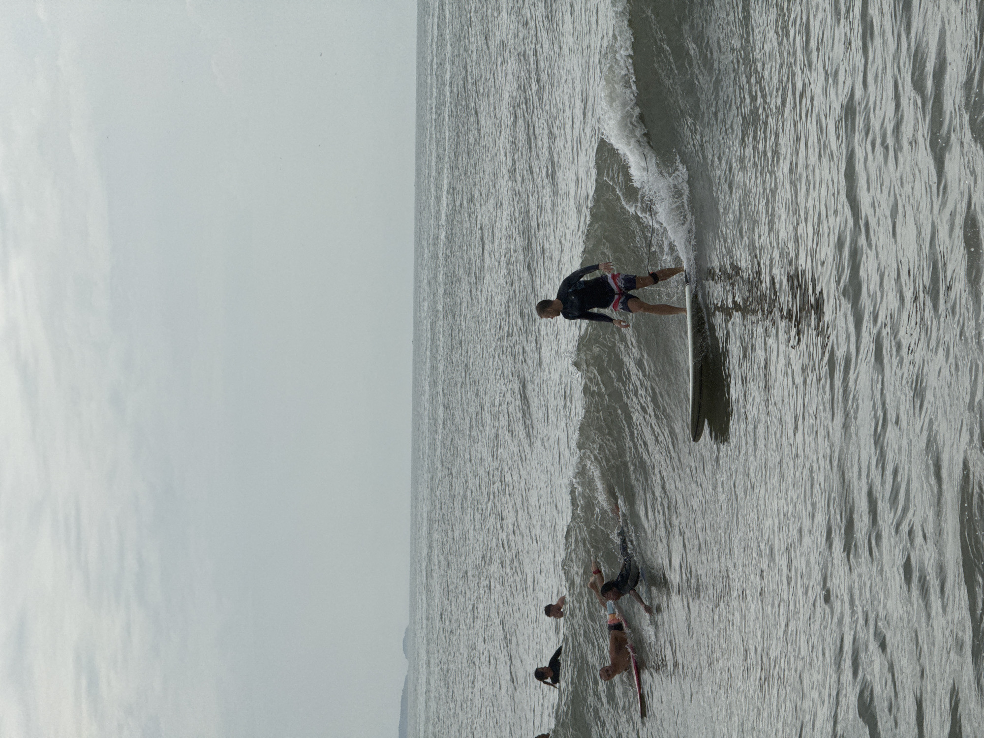 In Phan Thiết, Vietnam, several surfers are in the ocean on a gray, overcast day. One surfer, wearing a dark long-sleeved rash guard and patterned shorts, is actively riding a wave on a surfboard. In the background, other surfers are either paddling on their boards or partially submerged in the water. The sea is choppy, with visible waves and whitewater. The horizon line is flat, with a hazy sky above the ocean. A distant shoreline or landmass is visible on the left side of the image.