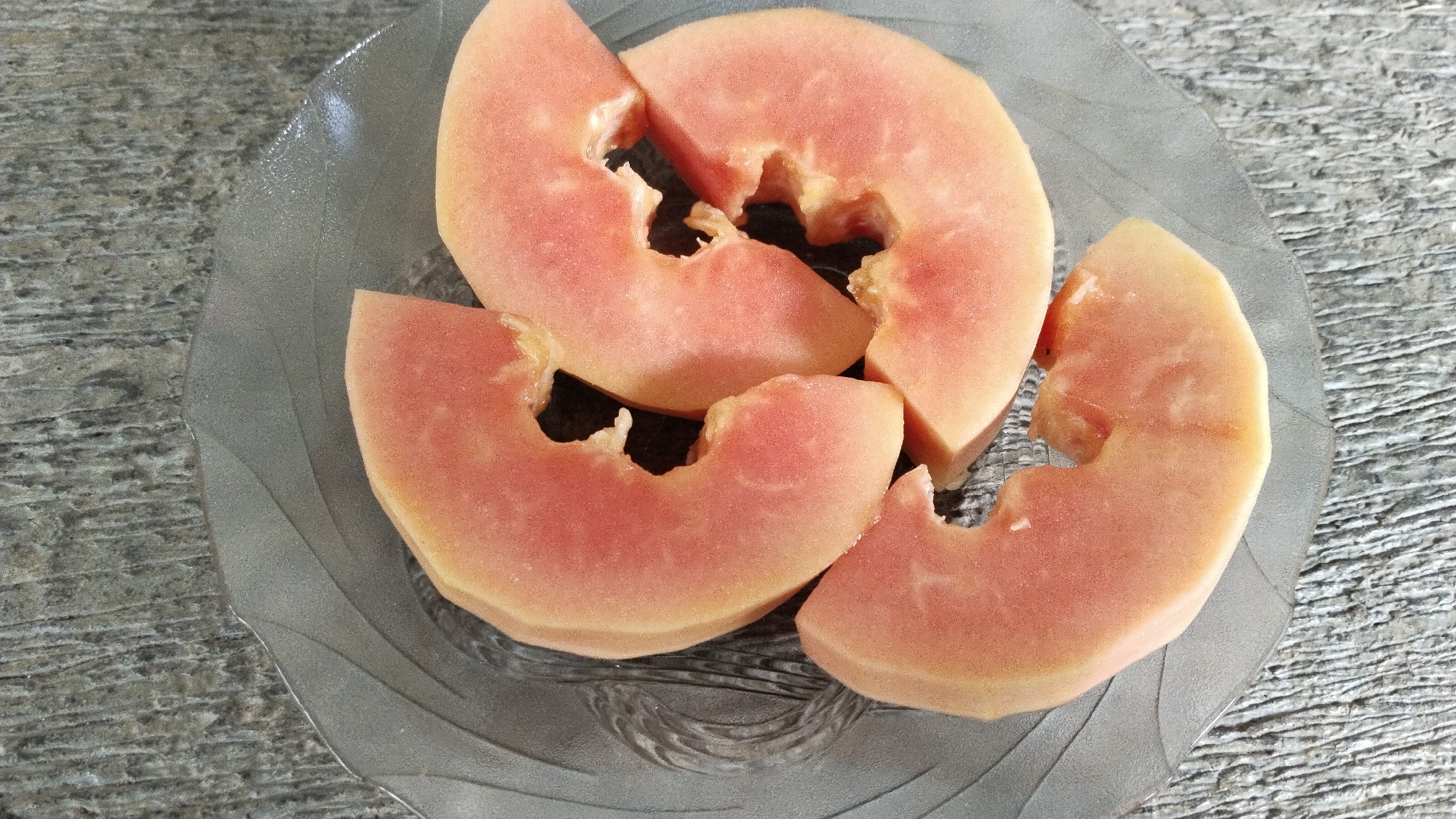 A clear glass plate containing three slices of guava sits on a gray, textured surface, likely a wooden table or concrete. The guava slices are roughly triangular, with some of the seed cavity visible. The flesh of the guava is a light pinkish-orange color. The plate has an embossed design with wavy lines. The background is a neutral gray texture. The lighting suggests an outdoor or well-lit indoor environment, with subtle shadows cast by the guava. The scene is static, with no activity or people present. The location is Margatunggal, Indonesia.