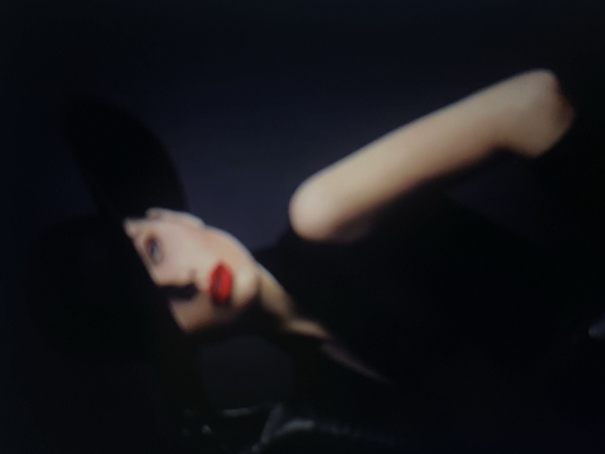 The image is a dimly lit, out-of-focus portrait of a woman wearing a wide-brimmed black hat and red lipstick. Her face is partially obscured by the shadow of the hat, and she is looking to the right. She is wearing a dark garment, possibly a dress or a jacket. The background is dark and indistinct, suggesting an indoor setting or a nighttime scene. There is no identifiable information that would link this image to the city of Kołobrzeg, Poland.