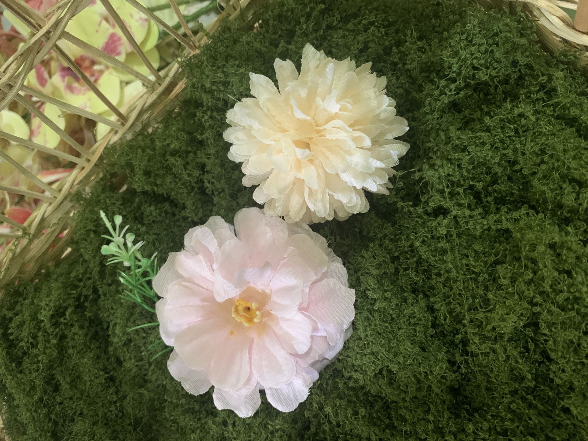Two artificial flowers, one light pink and the other a pale yellow, are placed on a bed of artificial green moss. The pink flower is larger and more open, with delicate, layered petals. The yellow flower is smaller and denser, resembling a chrysanthemum. To the upper left of the pink flower, a small sprig of artificial green leaves is visible. In the background, a woven basket with a light brown hue is partially visible, containing some blurry, colorful flowers with yellow and pinkish-red accents. The scene is lit from above, casting soft shadows. The location is not discernible from the image.