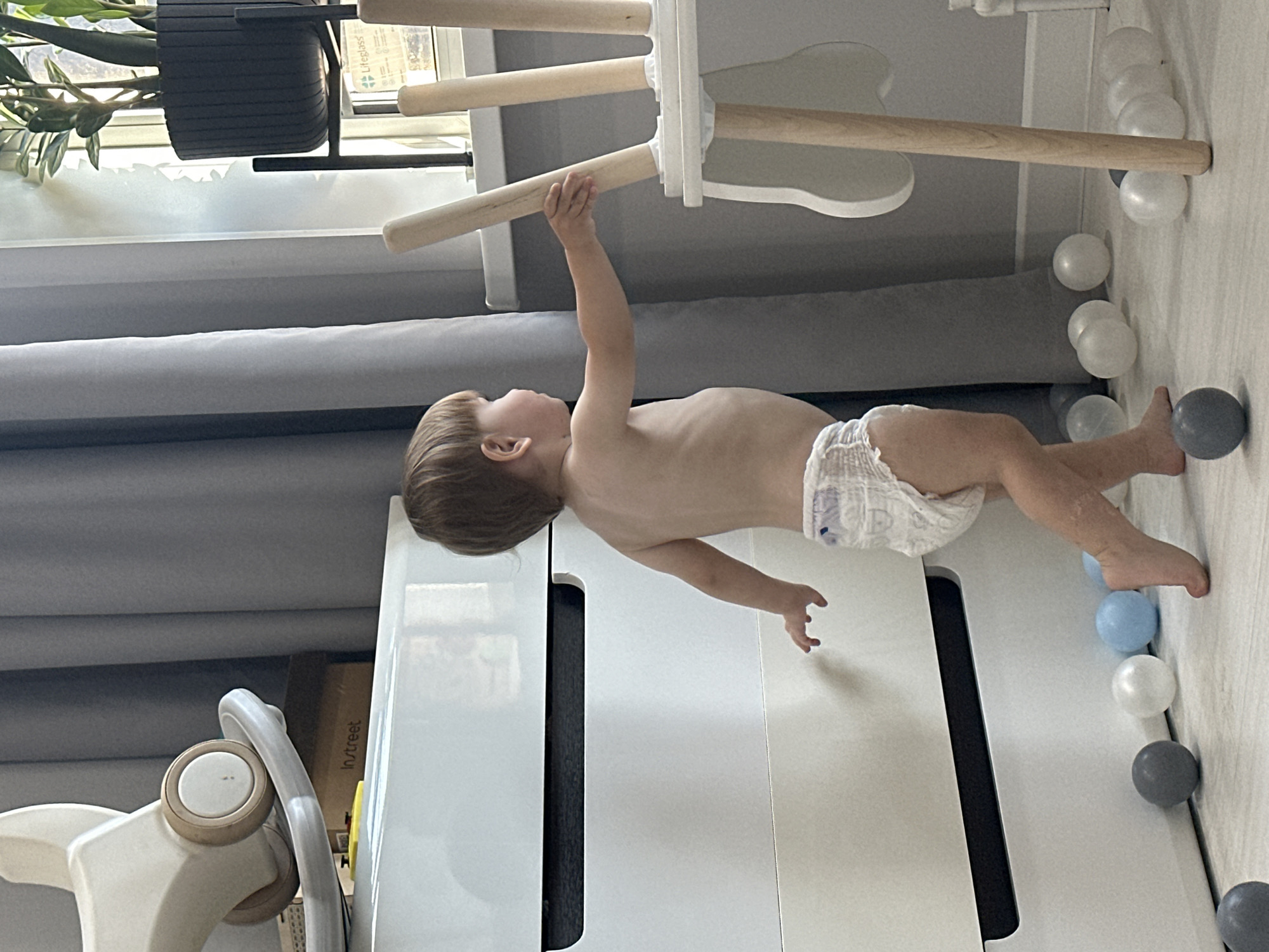 The image shows a toddler with brown hair, wearing a diaper, hanging from a wooden bar of a wooden indoor climbing structure. The structure has white frames and multiple wooden bars. The toddler's body is positioned horizontally, with their right arm extended upwards and grasping the bar. Their left arm is bent, and their legs are slightly bent. The toddler appears to be looking to the left, with their head turned.
The setting is indoors, likely a playroom or nursery. The floor is a light-colored wood or laminate. Scattered on the floor around the climbing structure are several small plastic balls in white, gray, and light blue. In the background, there are gray curtains and what appears to be a white piece of furniture. To the left of the climbing structure, there is a black pot with a green plant.
The lighting suggests it is daytime, possibly morning or afternoon, with natural light coming from an unseen window. There are no strong indications of the weather or specific emotions. The image is taken from a slightly low angle, looking upwards at the toddler.
The text "in:street" is visible on a yellow label on a cardboard box in the background. The location context provided is Эстачи, Russia.