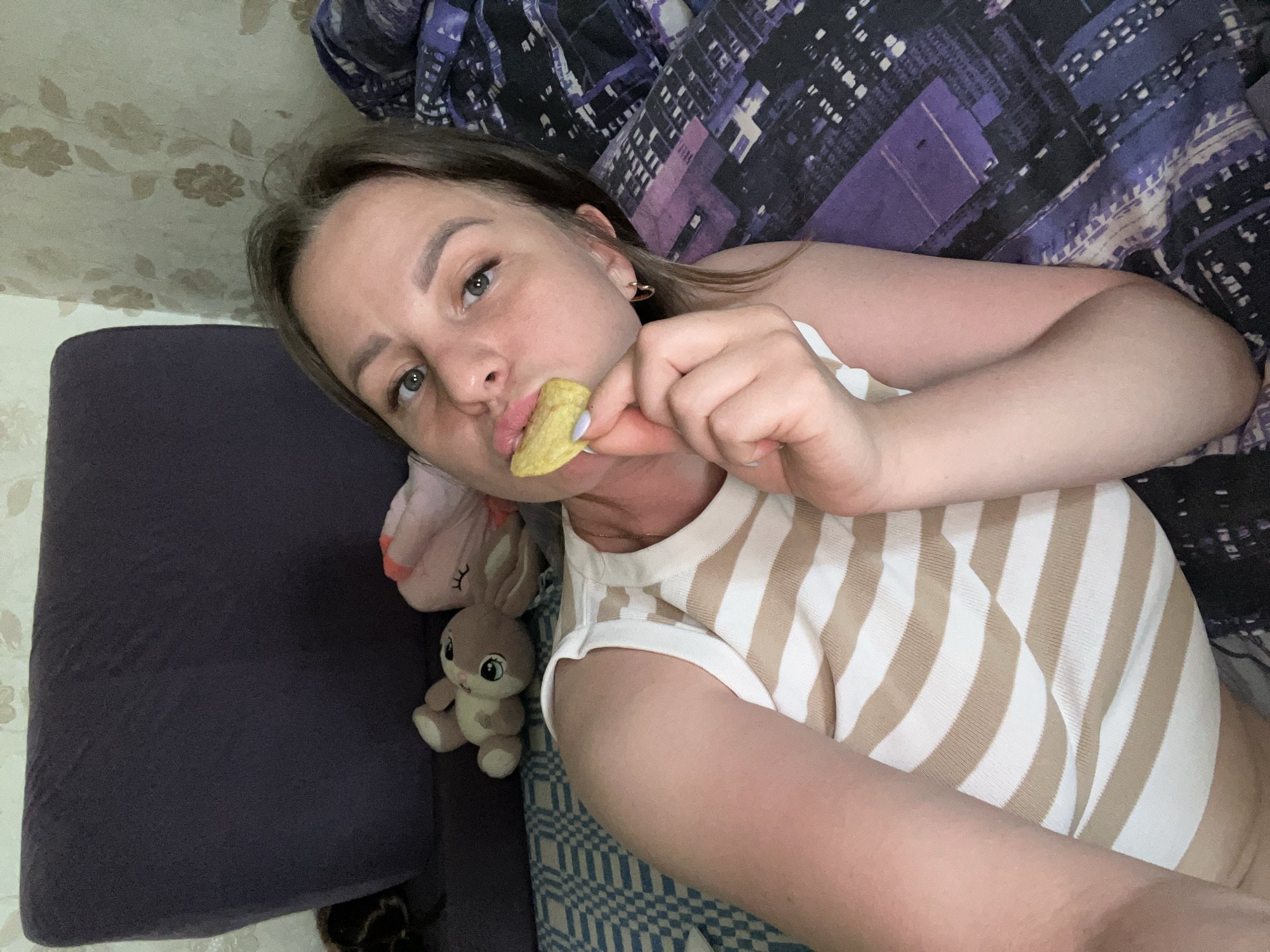 A woman is lying on a bed, eating a chip. She is wearing a striped shirt. A stuffed bunny and a pink unicorn are next to her. The background includes a patterned bedsheet and a wall with floral wallpaper. The location is unknown.