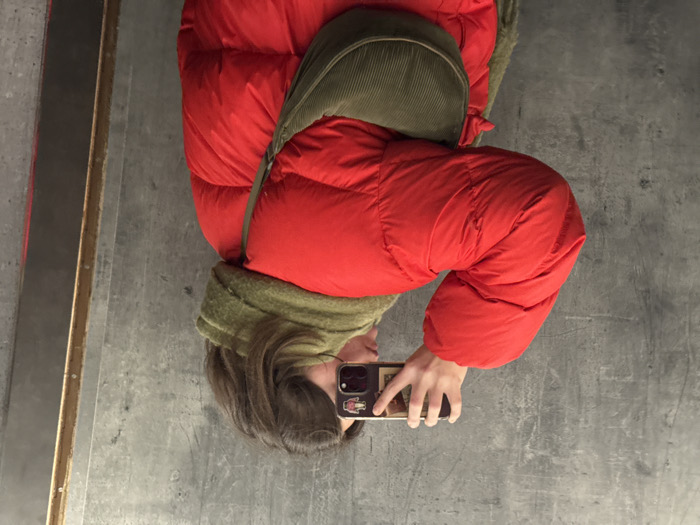 A person wearing a red puffer jacket with a green corduroy collar and bag is taking a selfie. They are holding a phone in their hand, and the phone case has stickers on it. The background is a grey concrete floor. A metallic vertical edge is visible in the left corner. The person's face is partially visible, showing part of their nose and mouth. Their hair is brown.