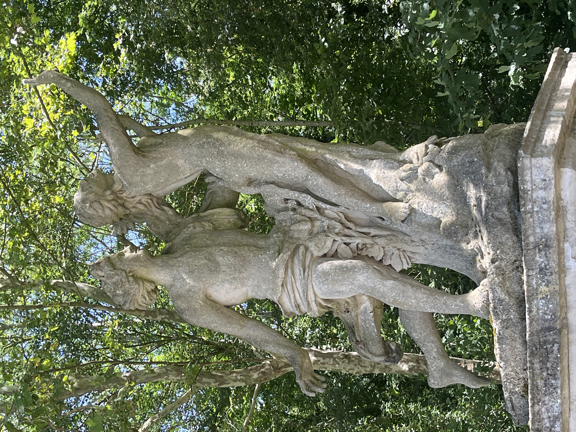This is a full, eye-level shot of a weathered stone sculpture of two nude figures, likely mythological, set against a backdrop of lush green trees and foliage under a bright, sunny sky. The sculpture is positioned on a stone pedestal in what appears to be a park or garden setting.
The sculpture depicts a male figure on the left, with his left leg extended forward, mid-stride. He is leaning back slightly, with his right arm raised and bent at the elbow, his hand reaching upwards. His body is draped with a cloth around his waist. To his right and slightly behind him is a female figure, her body angled away from the viewer. Her left arm is raised and extended upwards, and her right arm is bent, her hand resting on the male figure's shoulder or chest. Both figures are depicted with idealized, classical physiques.
The stone of the sculpture shows signs of age and weathering, with a patina of lichen and discoloration. The trees in the background are dense and green, with sunlight filtering through the leaves, creating dappled light and shadow on the sculpture and the surrounding environment. The overall impression is one of a serene and timeless scene.
The sculpture is located in Parc Frédéric Pic, in the city of Antony, France. This park is known for its collection of classical sculptures.
