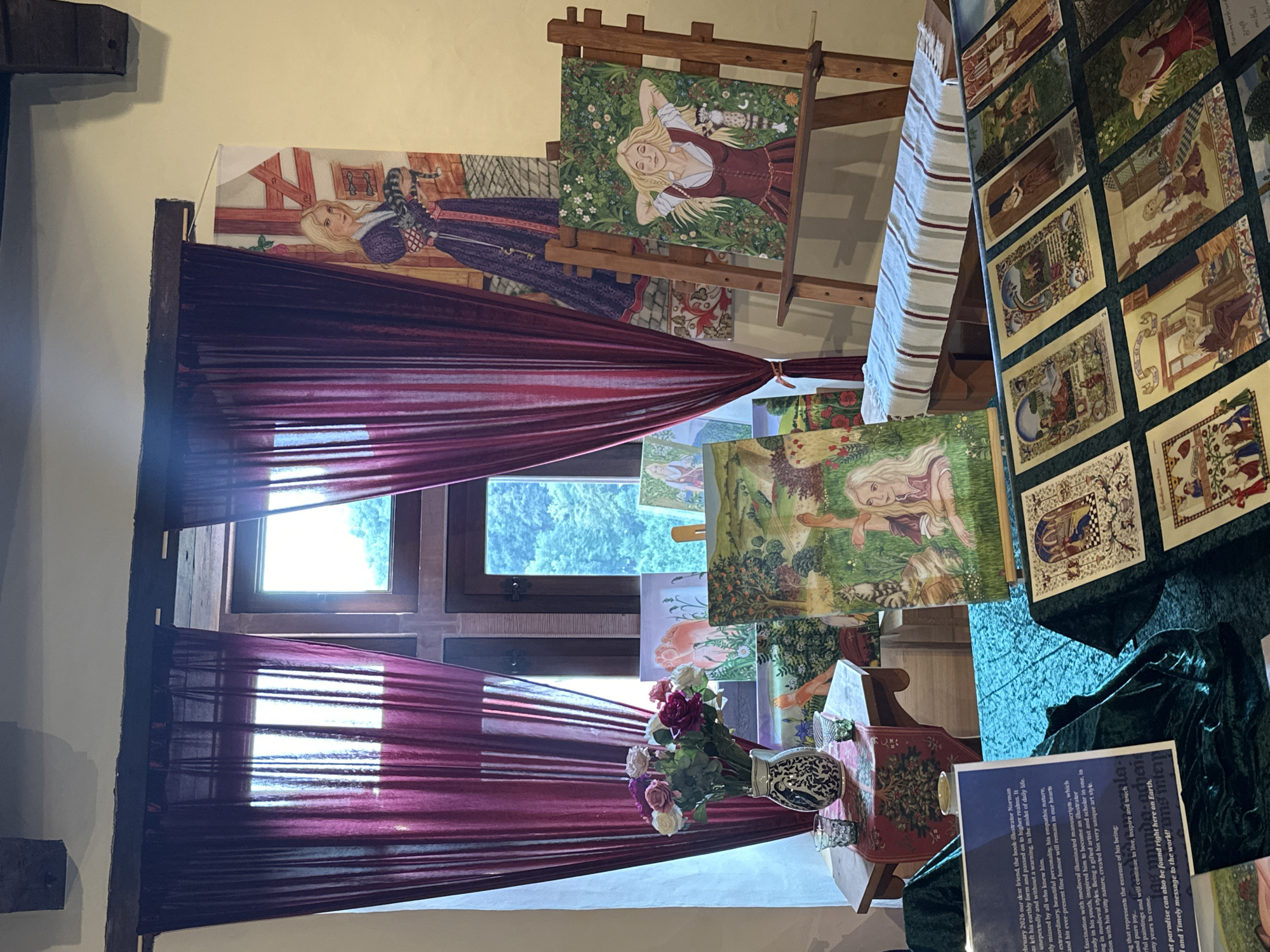 This image depicts an indoor exhibition space, likely a room within the Vianden Castle in Vianden, Luxembourg, given the style of the interior and the presence of historical-themed artwork. The scene is set up as a display for paintings and art prints. In the foreground, a dark green velvet cloth covers a table displaying a grid of rectangular art prints with detailed illustrations, possibly tarot cards or historical scenes. To the left, a rectangular blue plaque with white text provides information about an artist named Norman. Behind this, several paintings are arranged, including a vibrant landscape with poppies and a painting of a woman with blonde hair lying in a field of flowers, displayed on a wooden easel. Further back, a vase of artificial flowers sits on a hexagonal wooden table next to two small glasses. Large, sheer burgundy curtains hang from a dark wooden beam, partially obscuring two windows that offer a view of lush green foliage outside. To the right of the curtains, a large banner-style artwork hangs on the wall, featuring a woman with blonde hair holding a striped cat, dressed in a long purple robe. The overall ambiance is that of an art exhibition with a focus on historical or fantastical themes.