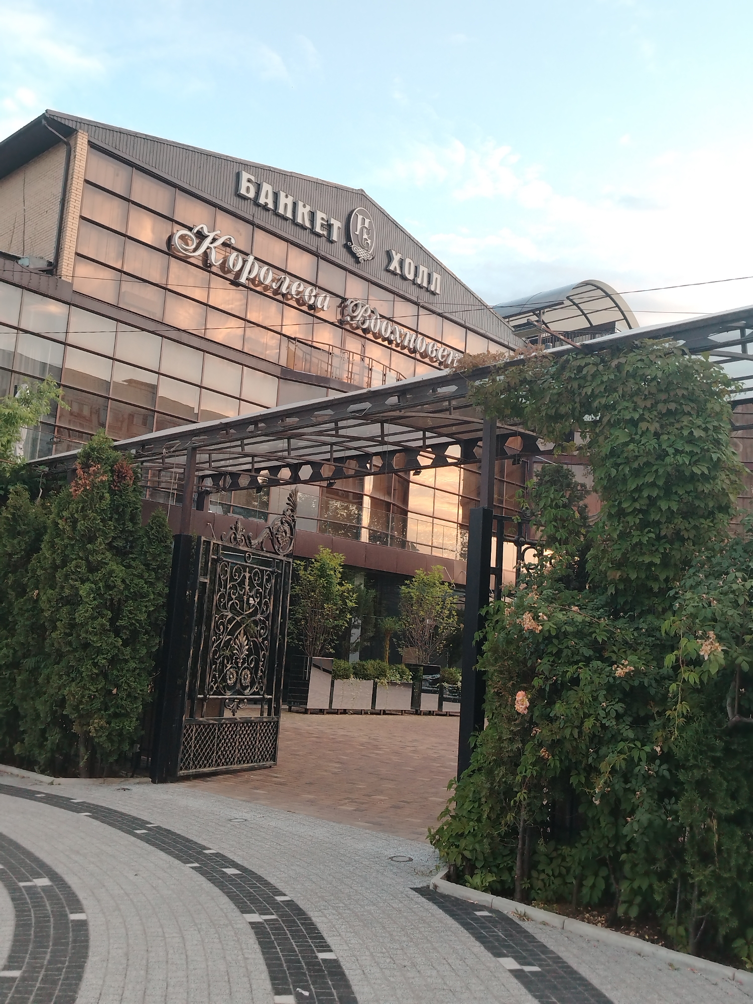 The image shows the exterior of a building labeled "БАНКЕТ ХОЛЛ" (Banquet Hall) with the name "Королева Вдохновения" (Queen of Inspiration) above the entrance. The building features a modern design with a glass facade that reflects the sky and surrounding environment. The entrance is marked by a decorative wrought-iron gate and lush green foliage, including trees and a large vine-covered structure. The foreground displays a paved pathway with a patterned design. The scene appears to be set during the day, with soft lighting and a clear sky. The location is identified as Krasnodar, Russia.