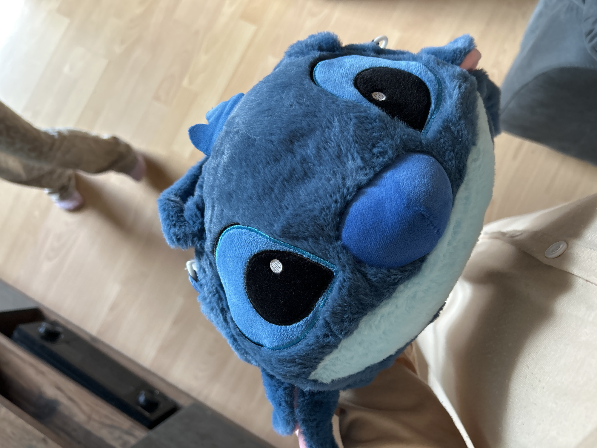 A close-up, eye-level shot shows a fuzzy blue plush toy, identified as Stitch from Disney's Lilo & Stitch, being held by a person wearing a tan shirt. The toy is positioned in the foreground, filling most of the frame with its large, black eyes and prominent blue nose. In the blurred background, a child wearing light brown pants and pink slippers walks across a light brown wooden floor. To the left, a dark brown wooden entertainment center with black hardware is partially visible. The scene appears to be indoors, likely in a residential setting within the city of Waldbröl, Germany.