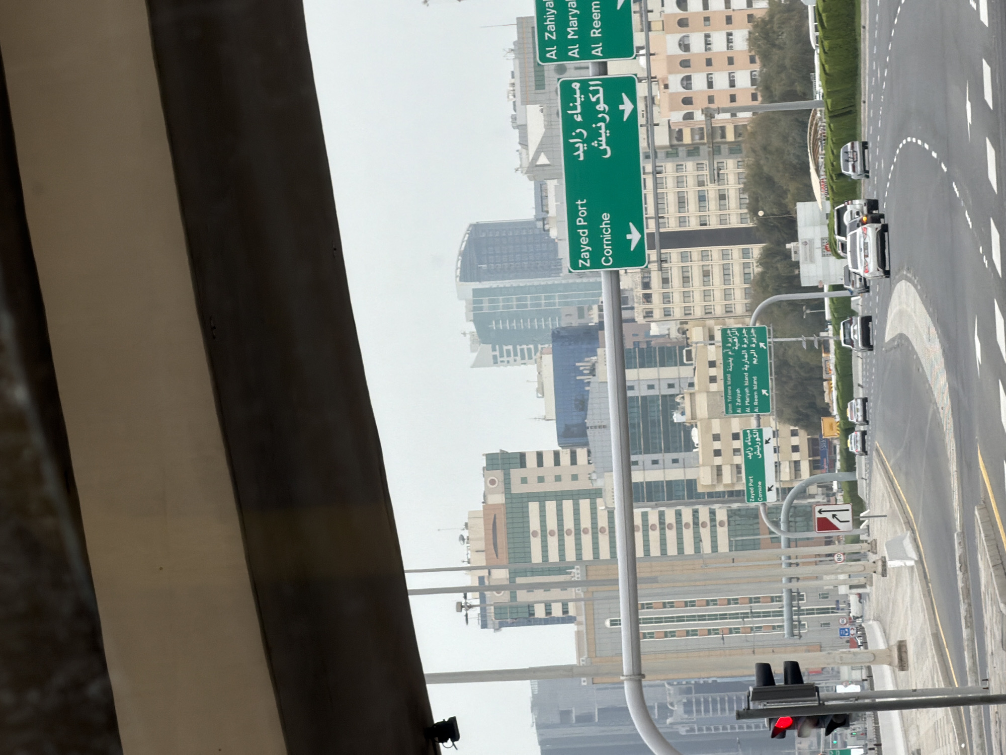 The image captures a street view in Abu Dhabi, United Arab Emirates, likely taken from within a vehicle. Dominating the scene are green directional road signs in both Arabic and English, indicating directions to Zayed Port and Corniche, as well as Al Zahiyah, Al Maryah, and Al Reem. Several high-rise buildings can be seen in the background. The road is visible, with cars driving, and there is a traffic light in the foreground with a red light.