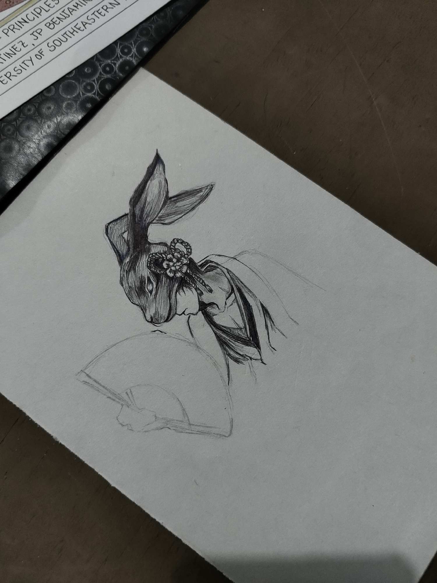 An overhead, angled shot captures a detailed pencil and ink drawing on a light grey paper, positioned on a dark brown, textured surface. The central subject is a partial depiction of a humanoid figure with the head of a rabbit. The rabbit's head, facing left, is rendered with visible cross-hatching for fur texture, featuring prominent ears and small, intense eyes. A stylized floral ornament with a braided knot adorns the side of the rabbit's head, appearing to be a hair accessory. The figure's torso is lightly sketched in pencil and partially inked, suggesting a traditional flowing garment with a high collar and wide sleeves. A hand, outlined in pencil, is holding an open hand fan, also in preliminary pencil sketch form, in the lower left portion of the drawing. To the upper left, a black notebook with a repetitive circular pattern is visible, topped by a white paper containing typed text snippets such as "PRINCIPLES," "MARTINEZ," "JP BENJAMIN," and "VERSITY OF SOUTHEASTERN." The location is unknown.