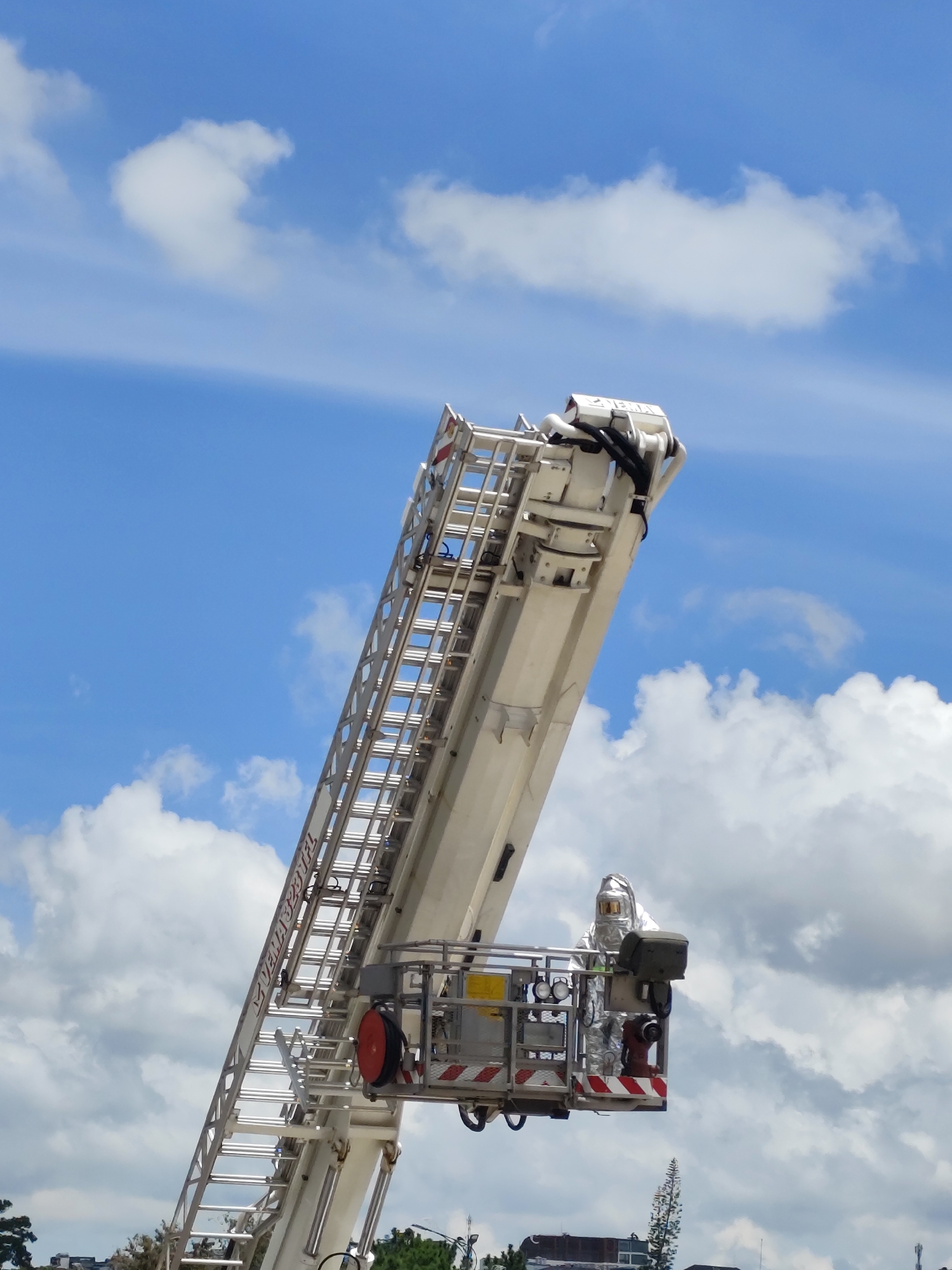 In Da Lat, Vietnam, a person clad in a reflective, silver hazmat suit stands in the basket of an extended fire ladder. The ladder, primarily white, angles upwards against a bright blue sky dotted with fluffy white clouds. The basket is equipped with safety railings and features red and white reflective tape along its edges. A red fire hose nozzle is visible within the basket, along with other equipment. The lower portion of the image shows a glimpse of buildings and trees, suggesting an urban or suburban setting.