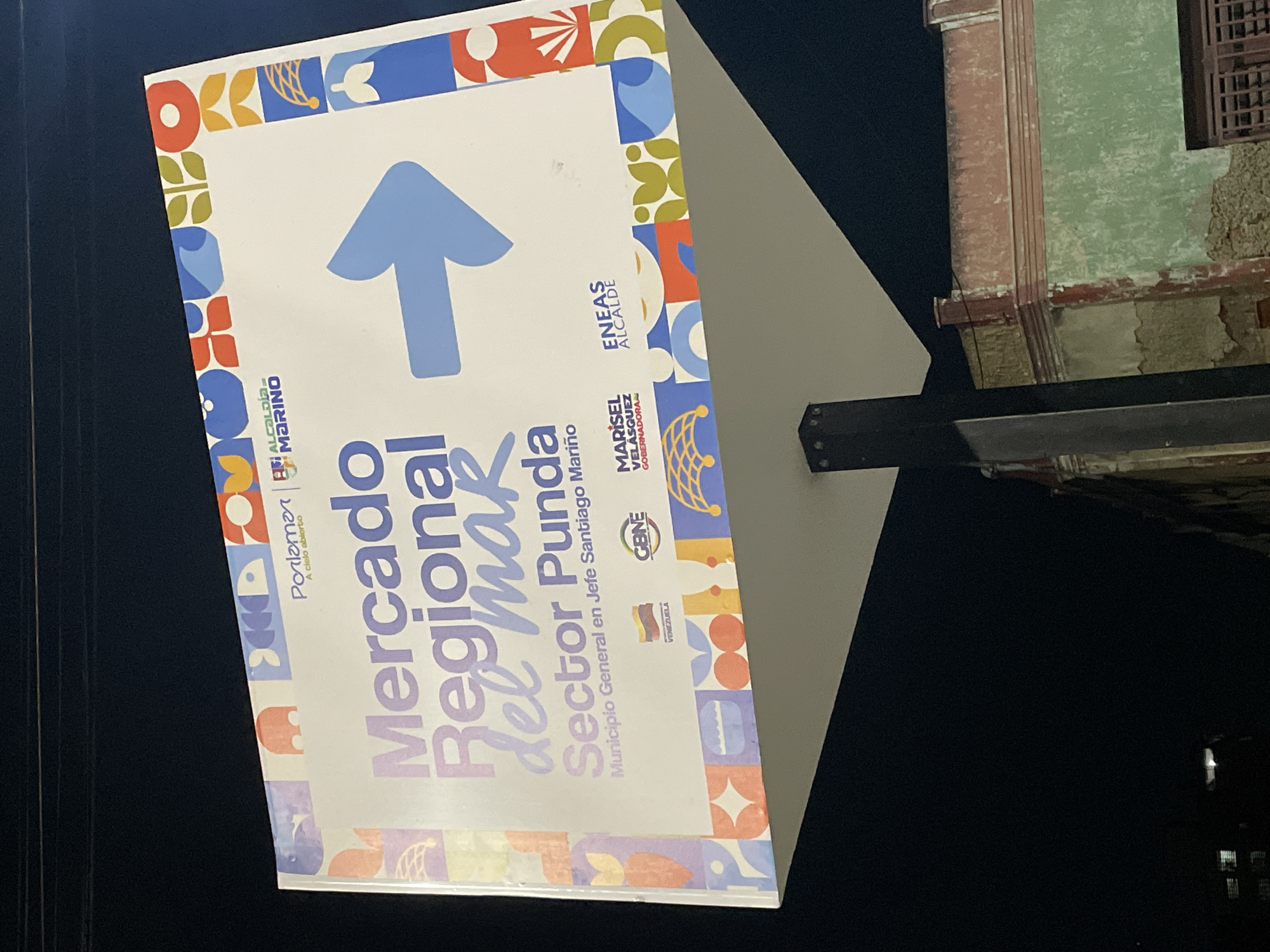 The image shows a street sign for the "Mercado Regional del Mar" (Regional Sea Market) in Sector Punda, Municipio General en Jefe Santiago Mariño, located in Porlamar, Venezuela. The sign features an upward pointing arrow, logos for various local government entities including the Gobernadora Marisel Velásquez and Alcalde Eneas, and colorful graphic elements around the edges. The sign is mounted on a metal pole. The background reveals a building with faded green paint and aged architectural details, suggesting an urban environment. The sky is dark.