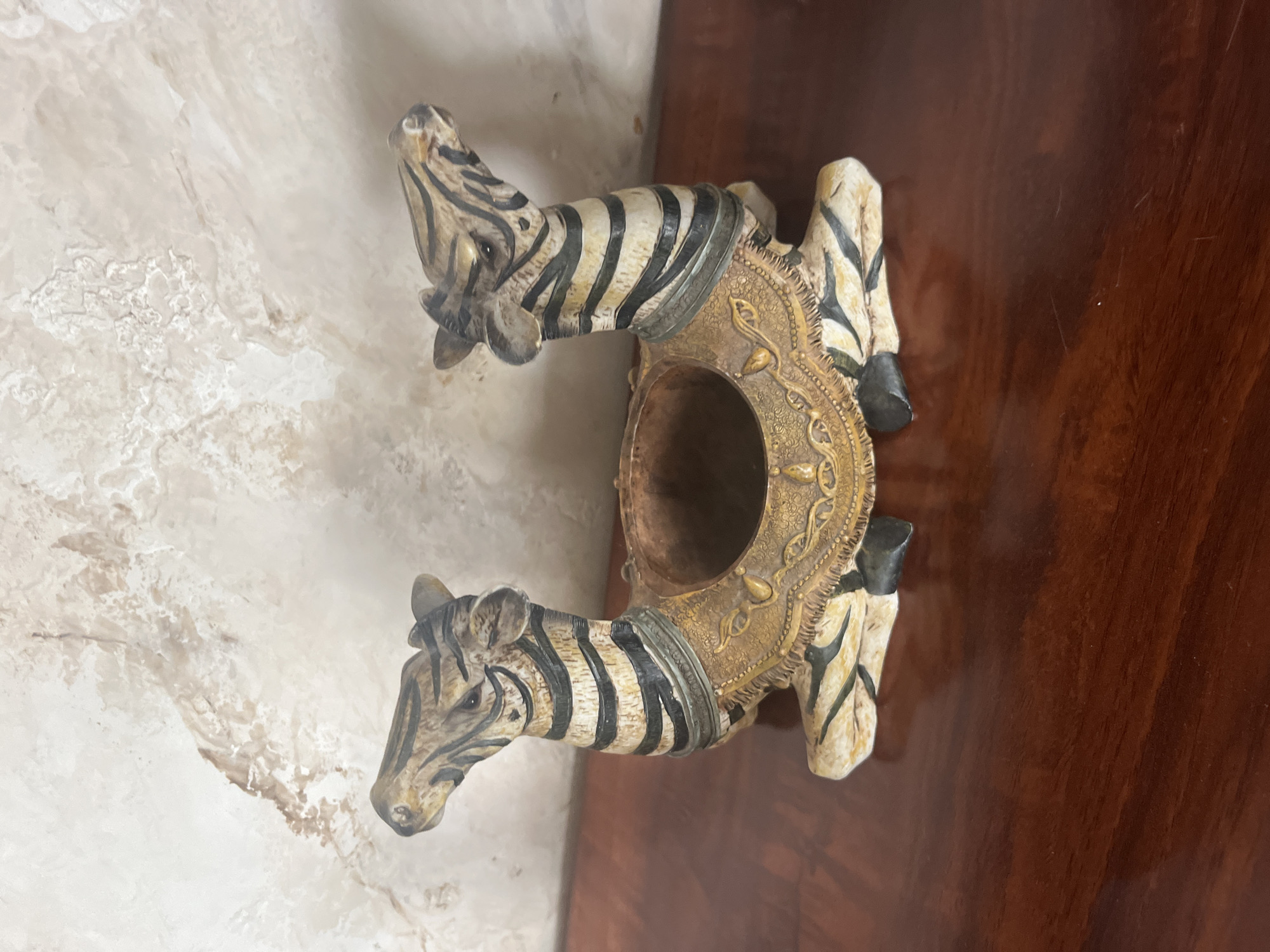 This is a medium shot, eye-level, indoor photograph of a decorative object featuring two stylized zebras. The object is positioned on a dark brown, polished wooden surface, likely a table or shelf. The background is a textured wall with a marble-like pattern in shades of beige and brown.

The decorative object is shaped like two kneeling zebras facing outwards, with their heads and necks curving upwards and inwards, creating a central space between them. The zebras are painted with black and white stripes, mimicking a zebra's coat, and have details painted in gold around their necks and bodies, suggesting elaborate adornment. The central space appears to be a hollow, possibly designed to hold something like a candle or a small decorative item.

The lighting is soft and diffused, casting faint shadows. The overall impression is of an ornamental piece displayed in a domestic setting. The image does not provide any information about the city of Vladimirovka, Russia, or any people, actions, or interactions.