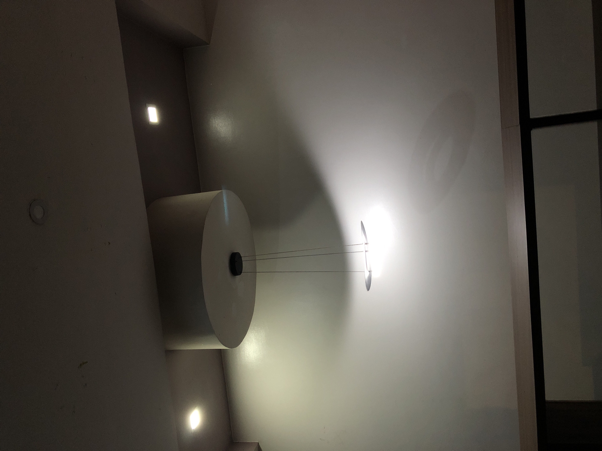 The image, captured from a low angle and rotated clockwise, depicts an indoor ceiling and adjacent wall segment illuminated by artificial light. The main subjects are several modern light fixtures. On the left side, a large, light-colored cylindrical object, possibly a decorative element or a light fixture enclosure, hangs from the ceiling.
To the right of this cylinder, a minimalist pendant light fixture extends from the ceiling via three slender wires. This fixture, appearing as a thin, possibly reflective or disc-shaped element, emits a bright, intense white light that casts significant illumination and shadows across the ceiling.
Additionally, two smaller, rectangular recessed lights are visible on the left portion of the ceiling or upper wall, emitting a warmer, yellowish glow. The ceiling surface itself is a light, neutral color, showing reflections and various shadows cast by the fixtures. On the far right edge of the frame, a dark, angular structure, possibly a doorframe, window frame, or a structural beam, provides a stark contrast to the illuminated ceiling.
There are no people or specific activities taking place in the image. The artificial lighting suggests an indoor setting, likely during the evening or in a space where natural light is not prominent. No text is visible. The overall impression is of a contemporary interior design focused on unique lighting elements.