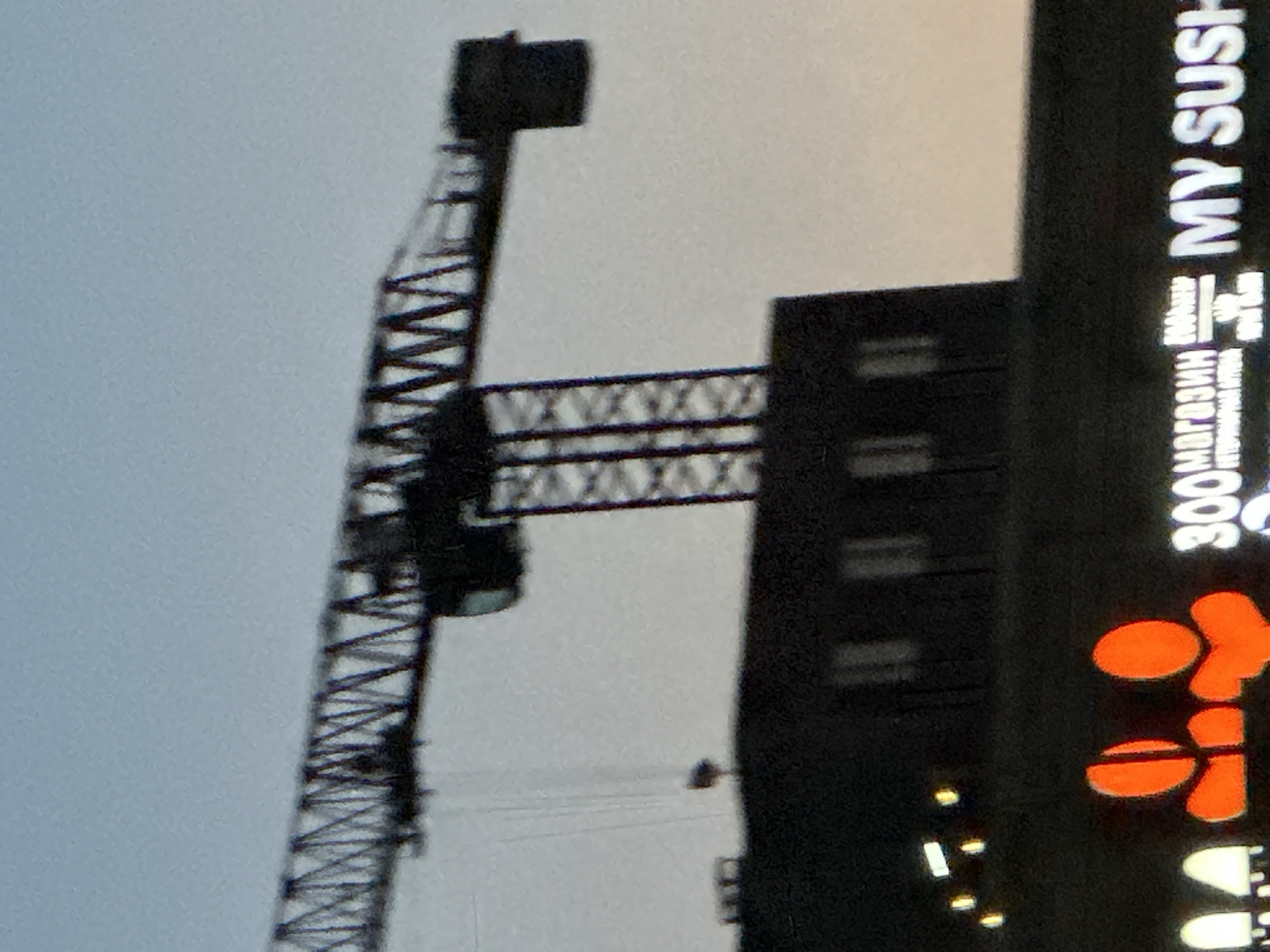 The image displays an outdoor scene dominated by a large construction crane on the left and a building with illuminated signs on the right. The sky, visible in the background, is light and appears overcast or in twilight, suggesting either early morning, late evening, or a very cloudy day.
A prominent dark silhouette of a truss-structured construction crane occupies the left and central parts of the frame, extending both vertically and horizontally. Thin lines, possibly cables or wires, are visible below the crane's horizontal arm.
To the right, a dark, multi-story building stands. Its facade features several horizontal bands, possibly windows. A bright, vertical sign runs along the right edge of this building. The sign displays white text oriented vertically, clearly reading "MY SUSHI." Below this, in Cyrillic script, the text "300 магазин" (300 store) is discernible, along with other partial Cyrillic characters that appear to relate to a store or business. Further down the sign, there are illuminated orange shapes, which could be stylized characters or a logo, possibly associated with the "MY SUSHI" brand. Scattered small white and yellow lights are also visible at the base of the building's illuminated facade.
The scene suggests an urban or commercial area, potentially under development given the presence of the construction crane. The visible text indicates businesses, likely including a sushi restaurant and a "300 store." The location context provided for this image is Бараўлянскі сельскі Савет, Belarus.