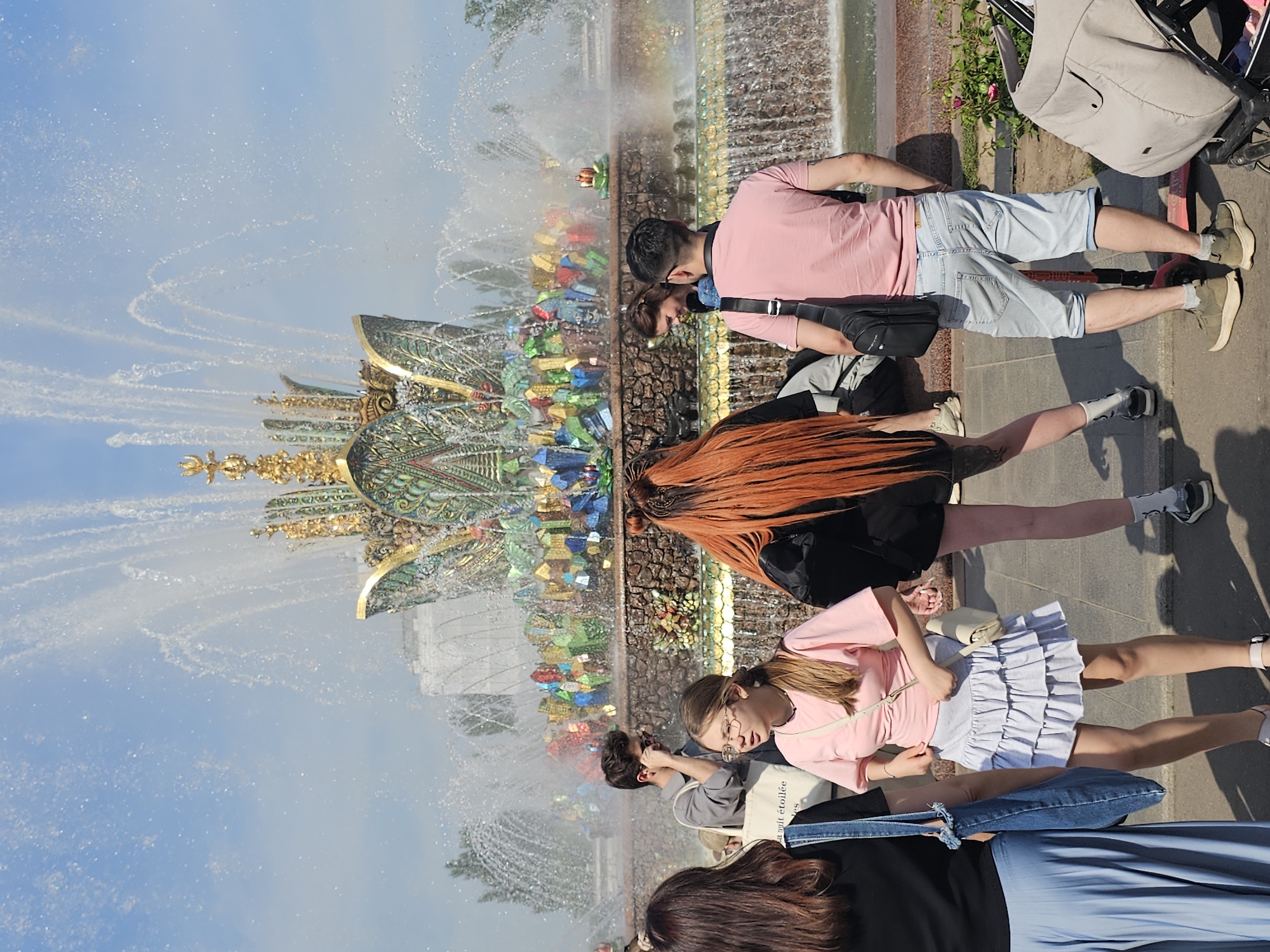 The image captures an outdoor scene at the "Friendship of Peoples" Fountain located within the VDNKh (Exhibition of Achievements of National Economy) complex in Moscow, Russia, under a clear blue sky. The prominent fountain features a golden, multi-tiered top with intricate green and multicolored mosaic-like patterns on its upper and middle sections. The lower tier displays a rough-textured base adorned with vibrant, irregularly shaped block-like decorations, from which water cascades into a basin. Multiple jets spray water upwards and outwards, creating a visible mist and a small rainbow effect to the right of the fountain.
In the left foreground, a person with dark brown hair is seen from the back, wearing a black top, a light blue pleated skirt, and carrying a blue denim-like tote bag. Adjacent to them, slightly right of center in the foreground, a young woman with shoulder-length light brown hair and glasses wears a pink short-sleeved top with a subtle front twist, a light grey ruffled mini-skirt, and a light-colored cross-body bag. She stands with a slight shift in weight. Further to the right in the foreground, a person with long, bright orange-red hair is visible from the back, wearing a black top, black shorts, white socks, and dark sneakers; tattoos are visible on their left thigh.
In the midground, to the left of the fountain, a young man with dark, curly hair and sunglasses pushed up on his head is wearing a grey short-sleeved collared shirt. He holds a white tote bag, partially obscuring text that includes "a nuit étoilée," and appears to be holding a phone to his ear. To the right of center in the midground, a man seen from the back wears a pink short-sleeved t-shirt, light blue denim capri shorts, green sneakers with light grey socks, and carries a black cross-body bag. Partially obscured behind him is a woman with dark hair, smiling and looking downwards.
On the far right, a beige baby stroller is partially visible. A small red kick scooter rests on the paved ground between the man in the pink shirt and the stroller. The ground is a light-colored paved surface, and some small green plants with red flowers are visible near the stroller. Green trees are present in the background, framing the fountain. The scene is brightly illuminated by natural sunlight.
