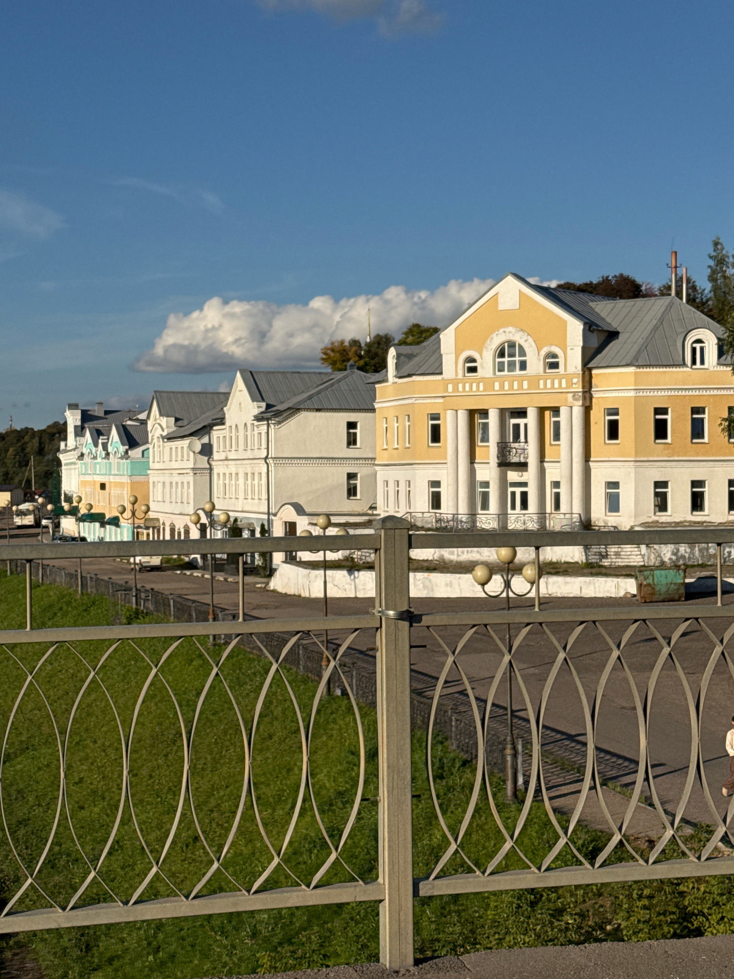In the city of Torzhok, Russia, along the Tvertsa River embankment, a row of historic buildings stands under a bright blue sky with scattered clouds. The buildings, varying in color from pale yellow to white and light blue, feature classic architectural elements such as arched windows, columns, and mansard roofs. A prominent yellow building with a grand entrance, supported by white columns, dominates the foreground of the building line. A decorative metal fence, with an intricate lattice design, runs along the embankment, separating a well-maintained green lawn from the paved walkway behind it. Several vintage-style lampposts line the walkway. A solitary figure, dressed in light-colored clothing, is walking on the far right of the frame along the embankment. The scene captures a serene day in this historic Russian town.