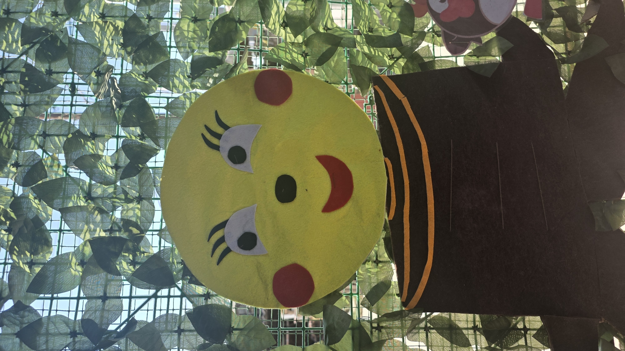 The image shows a close-up of a handmade bee decoration, possibly part of a larger display. The bee has a large yellow circular head with cartoonish features: two white oval eyes with black pupils, black eyelash-like lines above the eyes, a small black nose, two red circular cheeks, and a wide red smiling mouth. The head is attached to a dark brown body with horizontal orange stripes. The decoration is hung against a backdrop of green artificial leaves, which are attached to a green grid structure, suggesting an outdoor or semi-outdoor setting. A sliver of a light blue sky is visible through the leaves and grid. The background is slightly out of focus, drawing attention to the bee. The context of its location in Tokmok City, Kyrgyzstan, is not visually evident from the image itself; it is an external piece of information.