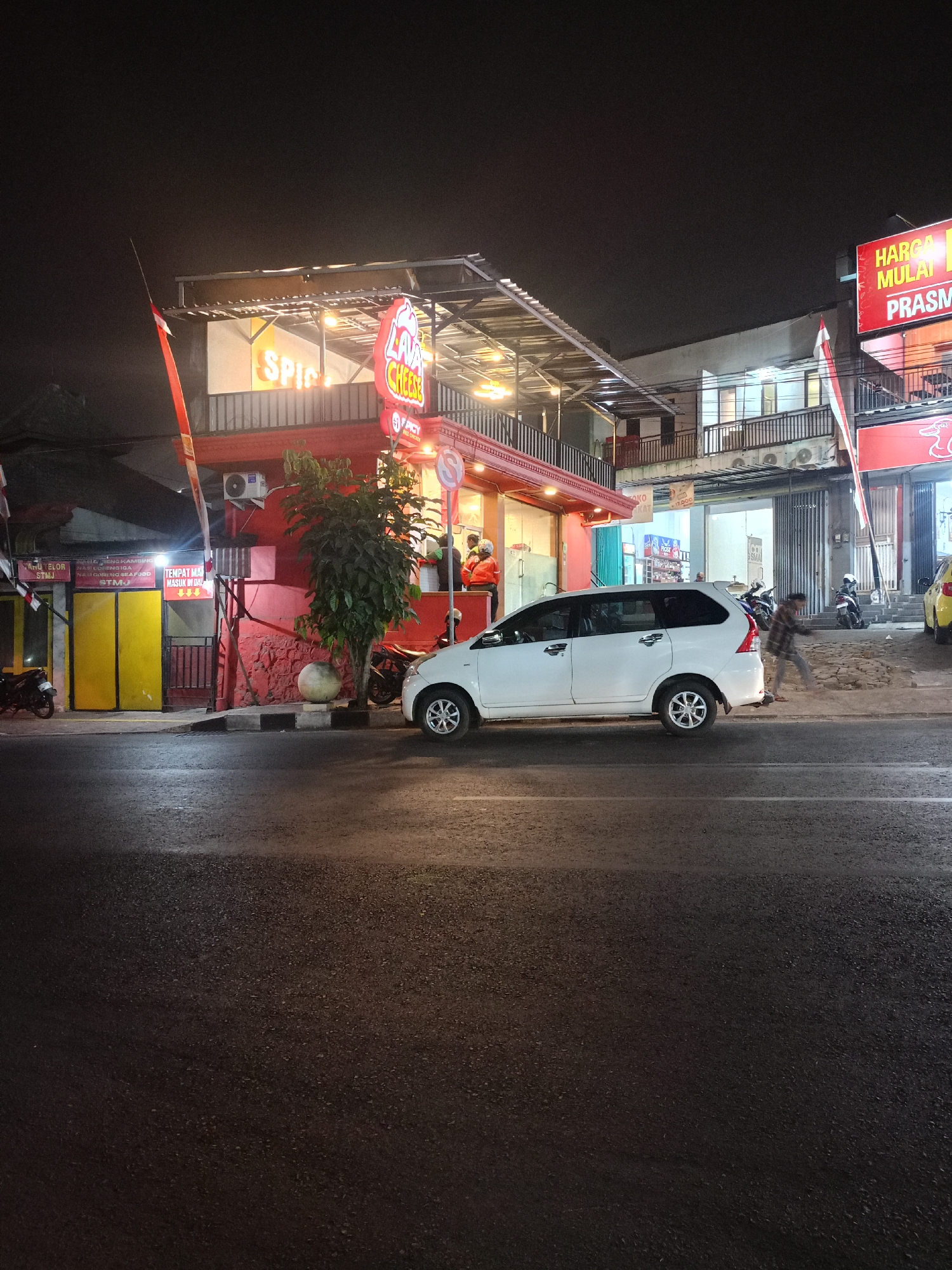 In the city of Batu, Indonesia, at night, a white minivan is parked on the side of a street in front of a brightly lit food establishment. The establishment has a red facade and a covered outdoor seating area with neon signs displaying "SPICY" and "LAVA CHEESE." Several people are visible inside and around the establishment, some appearing to be staff members wearing orange vests. Motorcycles are parked on the sidewalk. Across the street, another building with shops and apartments is visible, also illuminated at night. The street is paved and appears to be wet, reflecting the ambient light.