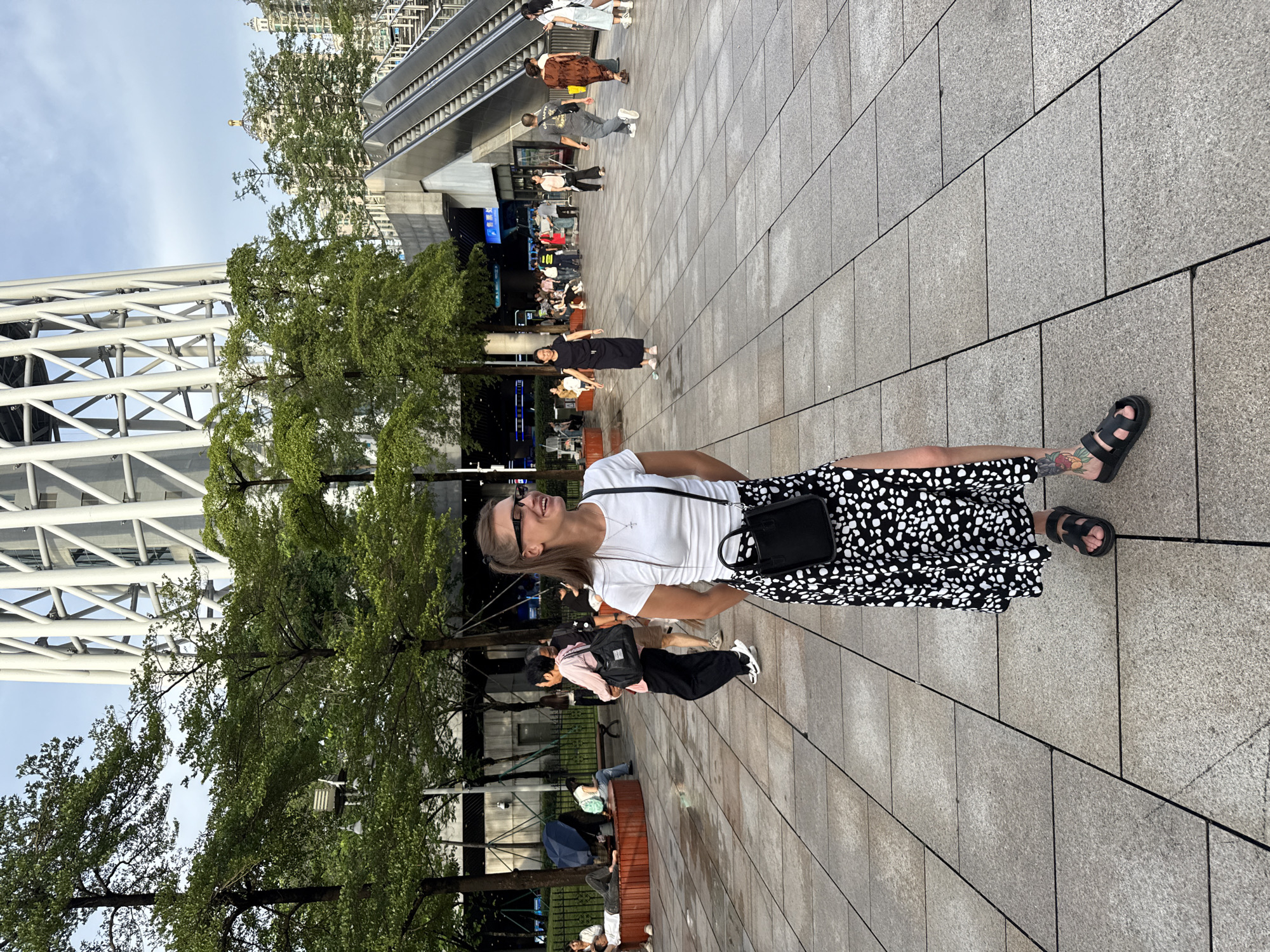 A young woman wearing glasses, a white t-shirt, and a black and white polka dot skirt with a thigh-high slit stands in an outdoor plaza. She has a black crossbody bag and black sandals. Behind her, other people are visible in the background, some sitting on benches, others walking. There are trees and a large, modern building with a distinctive white lattice structure in the background, likely the Canton Tower, located in Haizhu District, Guangzhou, China. Escalators are also visible to the right of the woman. The ground is paved with large, square tiles.