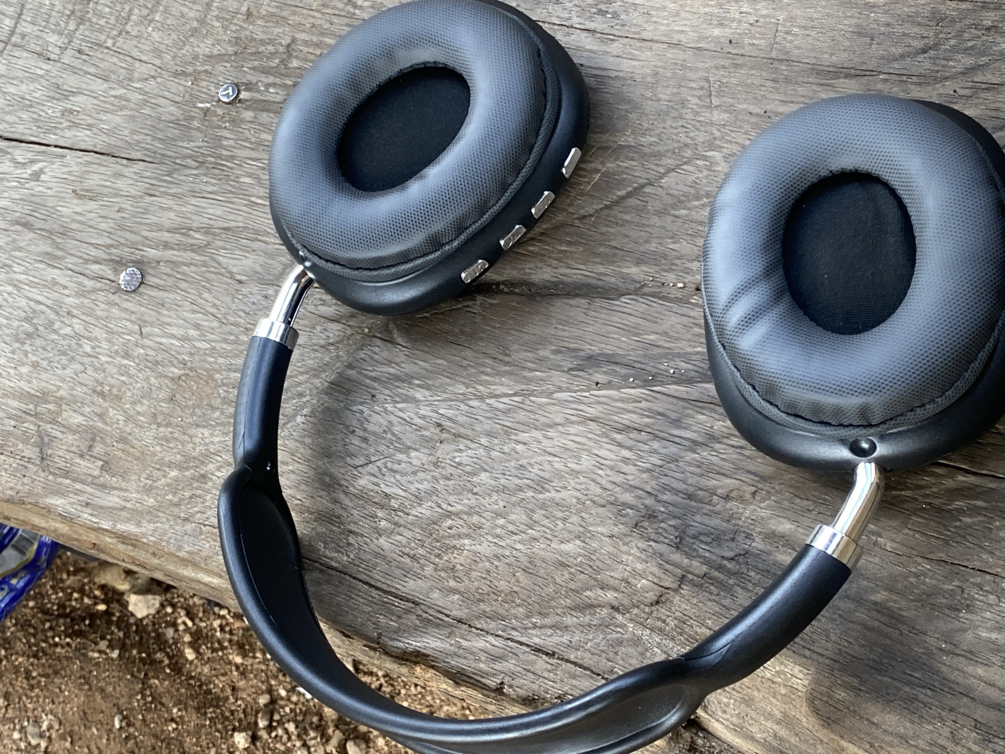 The image shows a pair of black over-ear headphones lying on a weathered wooden surface. The headphones are positioned with the headband curved, and the earcups are visible. The left earcup, partially obscured by the headband, has several small silver buttons along its side. The right earcup is more prominently displayed and also features a textured surface and silver buttons. The wooden surface appears to be a plank, with visible grain and some cracks. In the background, to the upper left, there is a patch of reddish-brown soil with small stones. The lighting suggests an outdoor setting. The scene is devoid of any human presence or activity.