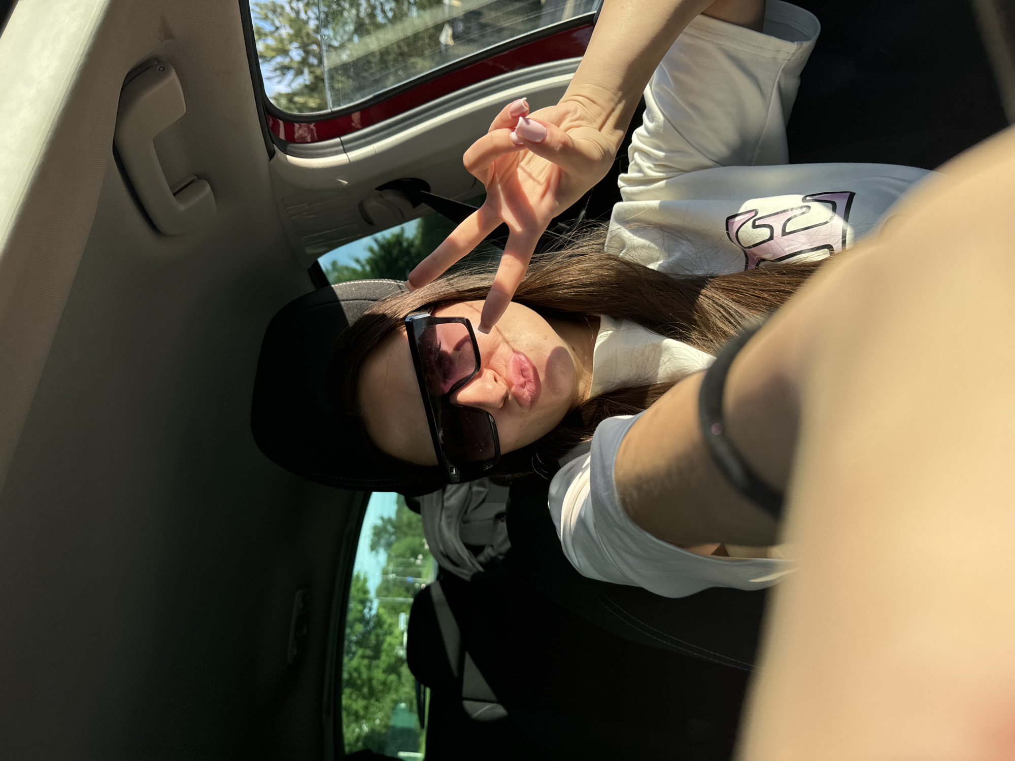 A young woman is taking a selfie from inside a car. She is wearing sunglasses and a white t-shirt with a graphic print. She is holding up a peace sign with her hand. The car is parked, with trees and sky visible through the windows. The location is unknown, including whether it is in Sharapovka, Russia.