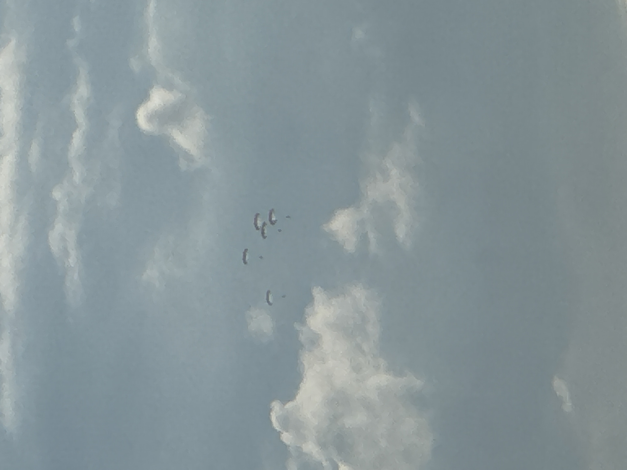 The image captures a sky filled with a mix of light and dark clouds. In the upper central portion of the frame, five paratroopers are descending. They are spread out, with four paratroopers clustered together and one slightly further to the right. The paratroopers are wearing dark-colored jumpsuits and their parachutes are open and visible as small, lighter shapes against the cloudy sky. There is no discernible location within the city of Krasnodar, Russia, as the image solely depicts the sky and clouds.