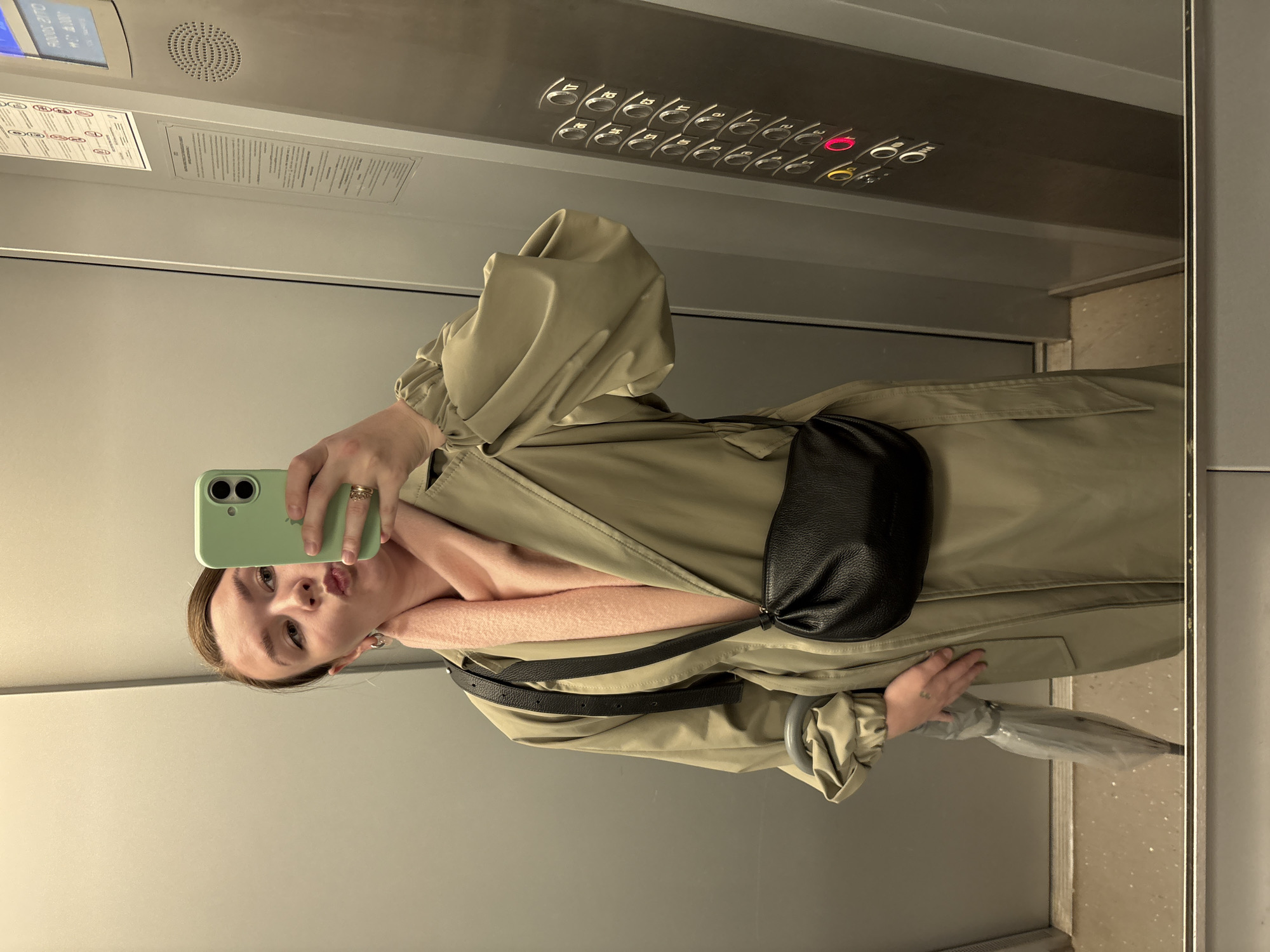 A woman takes a mirror selfie inside an elevator. She holds a light green smartphone, obscuring part of her face, and poses with a slight pout. She is dressed in a light olive green trench coat over a light pink scarf, and wears a black fanny pack or crossbody bag across her chest. A ring is visible on her finger. A clear, folded umbrella is partially visible, held in her left hand, suggesting possible rain or an expectation of it.
The elevator interior features reflective, light grey-beige walls. The control panel, visible in the mirror, shows a column of numbered buttons from 1 to 16. Several buttons are illuminated, specifically floors 6, 10, and 12 in red, and another button further down in yellow. A digital display screen and a circular speaker grille are also visible above the buttons. A notice board with multiple lines of unreadable text is affixed to the wall to the left of the control panel.
The scene depicts a casual, personal moment in an indoor setting with artificial lighting, likely within a building in Izhevsk, Russia.