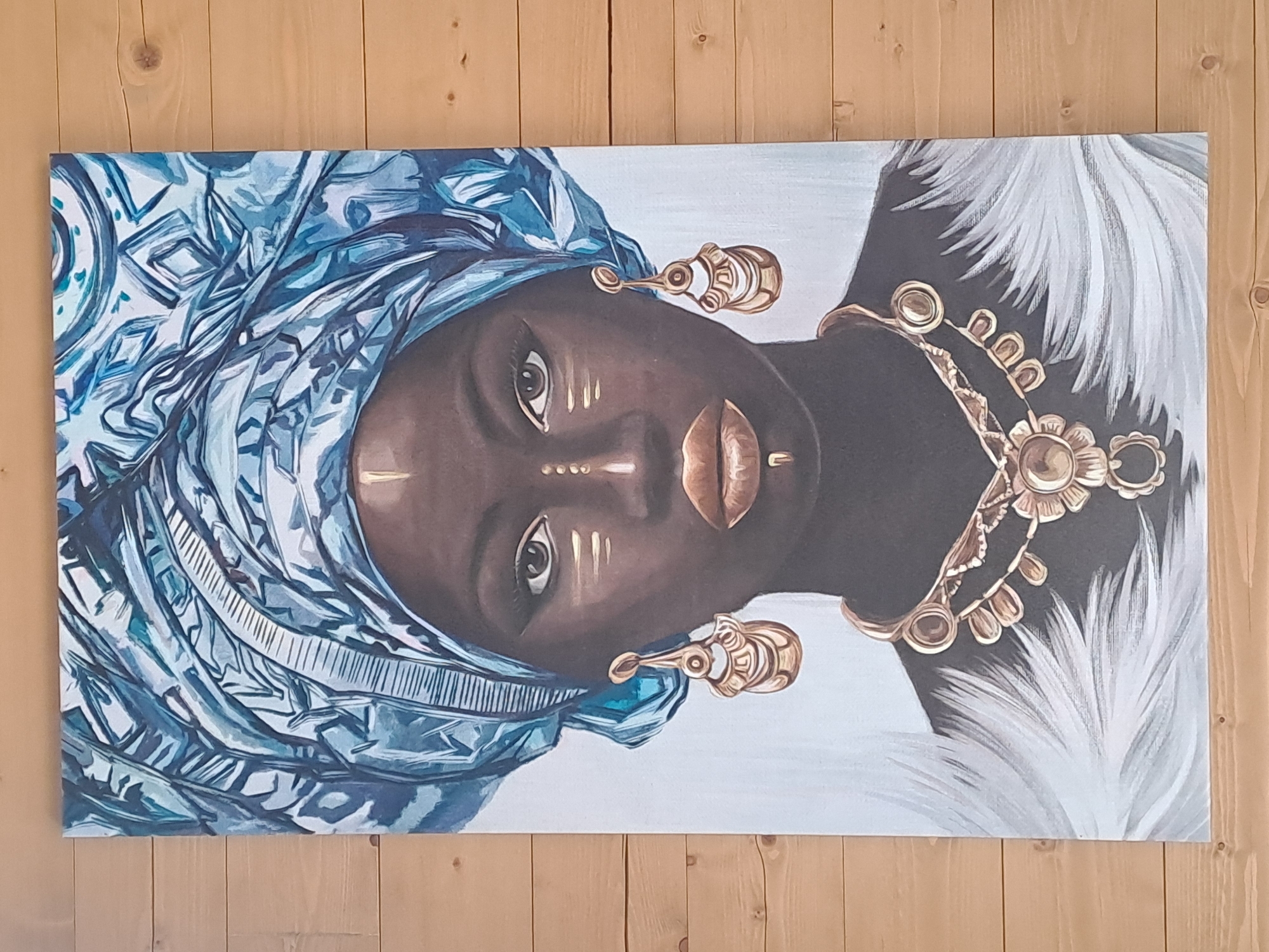 A vertical, digitally rendered painting of an African woman with dark skin is displayed on a wooden wall. The woman is adorned with a large blue headwrap, gold earrings, and a gold necklace. Her eyes are open and looking directly at the viewer. The background of the painting is a light blue and white gradient, with white feathery elements framing the bottom of the image. The wooden wall behind the painting consists of horizontal planks of varying light brown shades. The painting is centered on the wall, with roughly equal amounts of the wall visible on the left and right sides.
The location is the city of Krasnyi Ogorok, Russia.