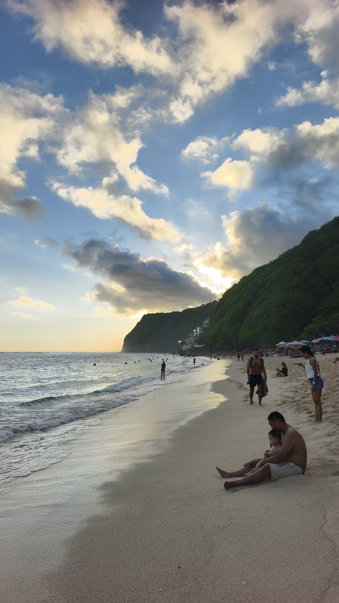 The image depicts a beach scene in Ungasan, Indonesia. The beach is sandy, with gentle waves lapping at the shore. Several people are scattered along the beach, including some swimming in the ocean, walking along the shoreline, or relaxing on the sand. In the foreground, a man sits on the sand with a young child in his lap. A woman in denim shorts and a white top is to the right of them. A man in black shorts is walking away from the water. The background features a lush, green hillside extending along the coast. A break in the hillside reveals a series of white buildings that look like small resorts. The sky is partly cloudy, with the sun creating a soft glow behind the clouds, suggesting it's either sunrise or sunset. The overall mood is peaceful and serene.