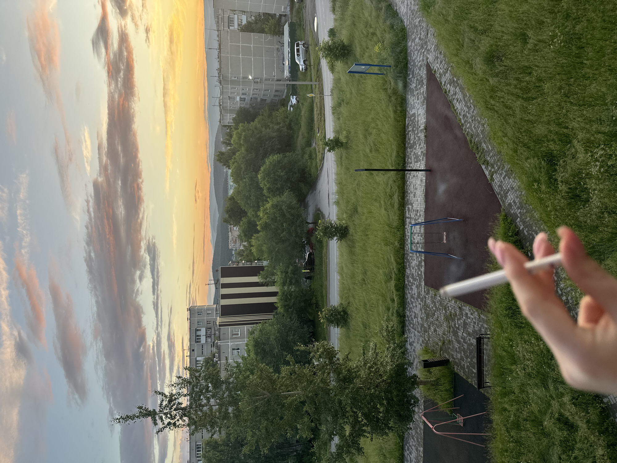 In Miass, Russia, at sunset, a person's hand, with pink-painted fingernails, holds a lit cigarette in the foreground. The background features a view of residential buildings, trees, and a playground. A road runs through the scene, with several cars parked or driving. The sky is filled with clouds, tinged with orange and pink from the setting sun. The overall environment appears to be a typical urban residential area.