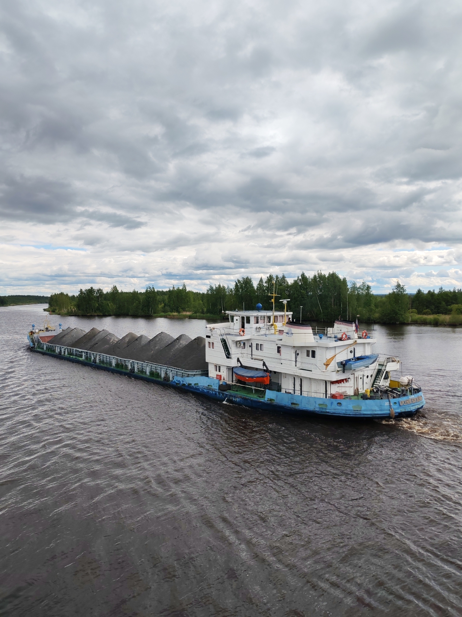 A barge carrying a load of dark gray material, possibly gravel or sand, is moving down a wide, calm river. The barge is long and flat, with a white superstructure at the rear containing multiple decks and windows. The superstructure is painted with blue trim and has life preservers visible on the exterior. The barge appears to be propelled by a tugboat, though only the rear of the tug is visible, creating a wake in the water.
The riverbanks on either side are lined with dense, green trees. The sky above is overcast with a dramatic mix of gray clouds, suggesting impending or recent rain. The overall atmosphere is one of industrial transport occurring in a natural, somewhat remote setting. The location is identified as Ozernoe Ust'e, Russia.