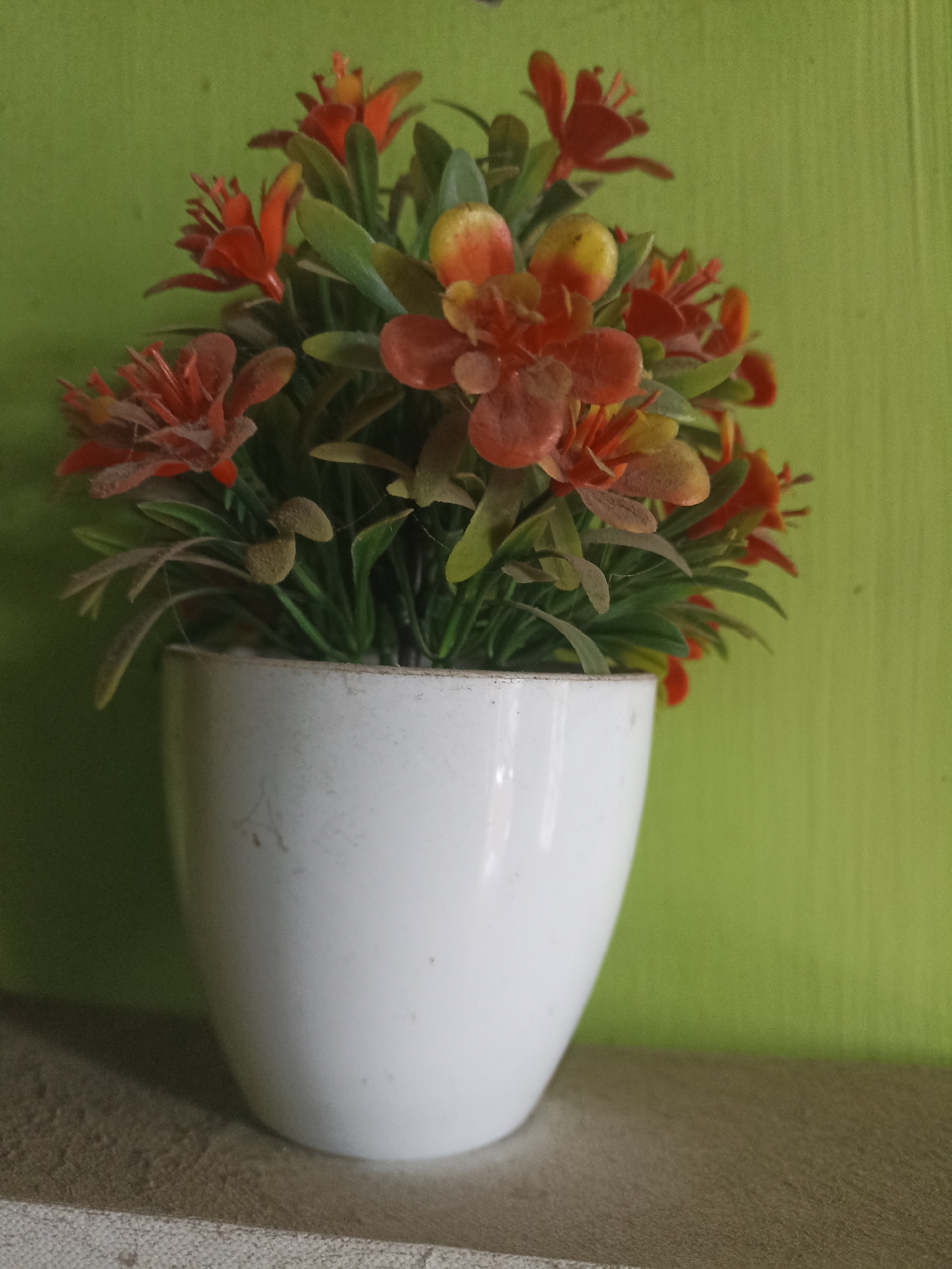 An artificial plant with orange and yellow flowers and green leaves is in a white pot on a textured surface. The background is a light green wall. The scene is indoors.