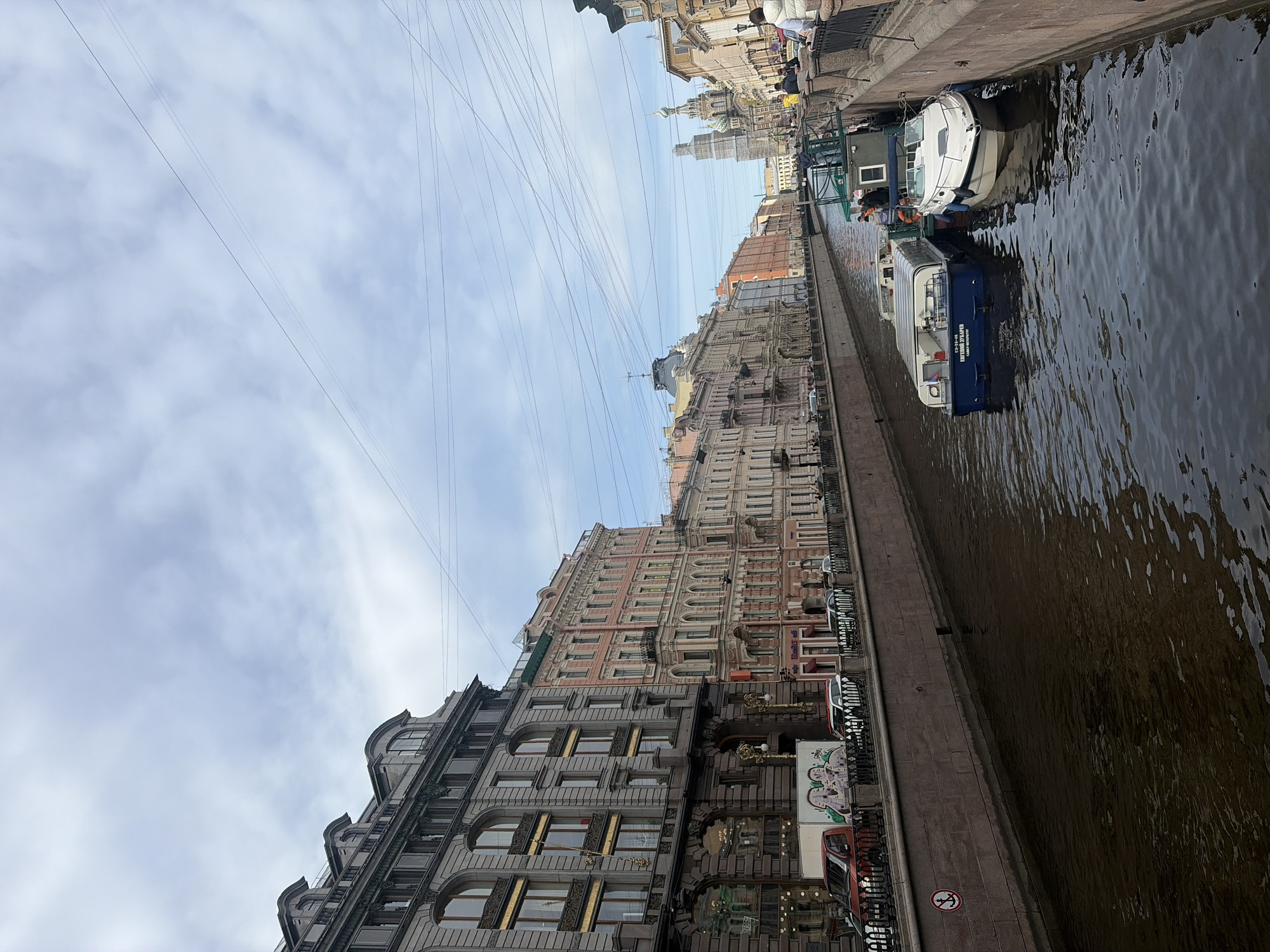This is a photograph taken in Saint Petersburg, Russia, showing a canal lined with buildings on the left and water with boats on the right. The buildings are mostly 4-5 stories tall, with varied architectural styles and colors, from grey stone to pink brick. Power lines stretch across the sky above the buildings. A stone embankment runs along the water's edge, with cars and railings visible. In the canal, several boats are docked. In the distance, the top of the Church of the Savior on Spilled Blood can be seen. The sky is cloudy.