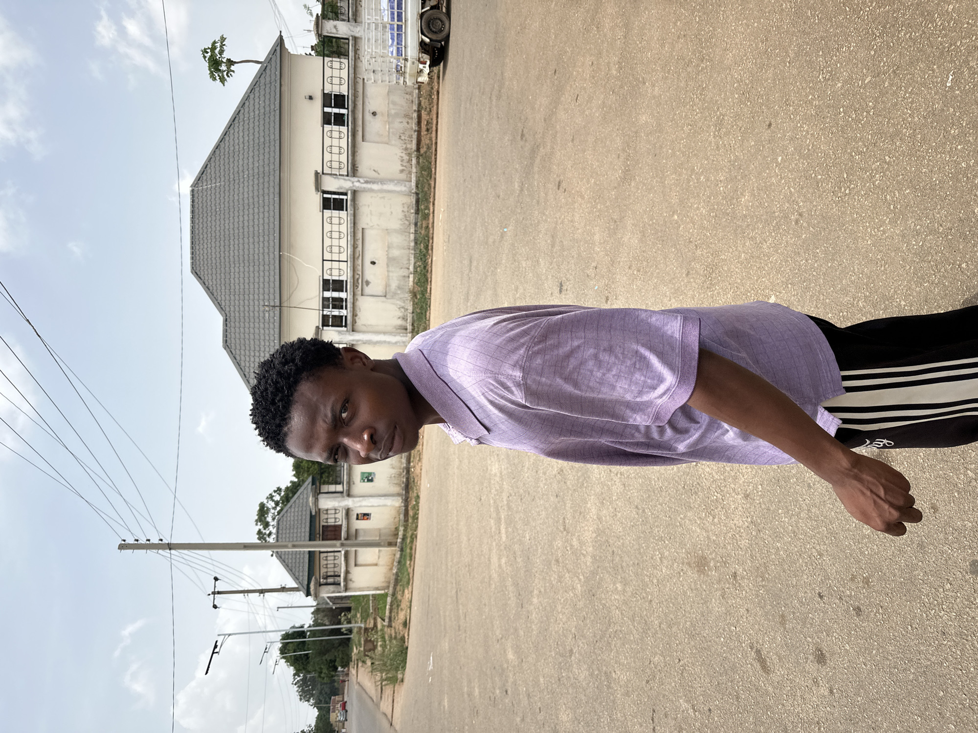 A young man stands on a paved road in Bukuru, Nigeria, looking directly at the camera. He is wearing a light purple collared shirt and black pants with white stripes down the side. The road appears to be a residential street, with buildings and power lines visible in the background. The sky is overcast.