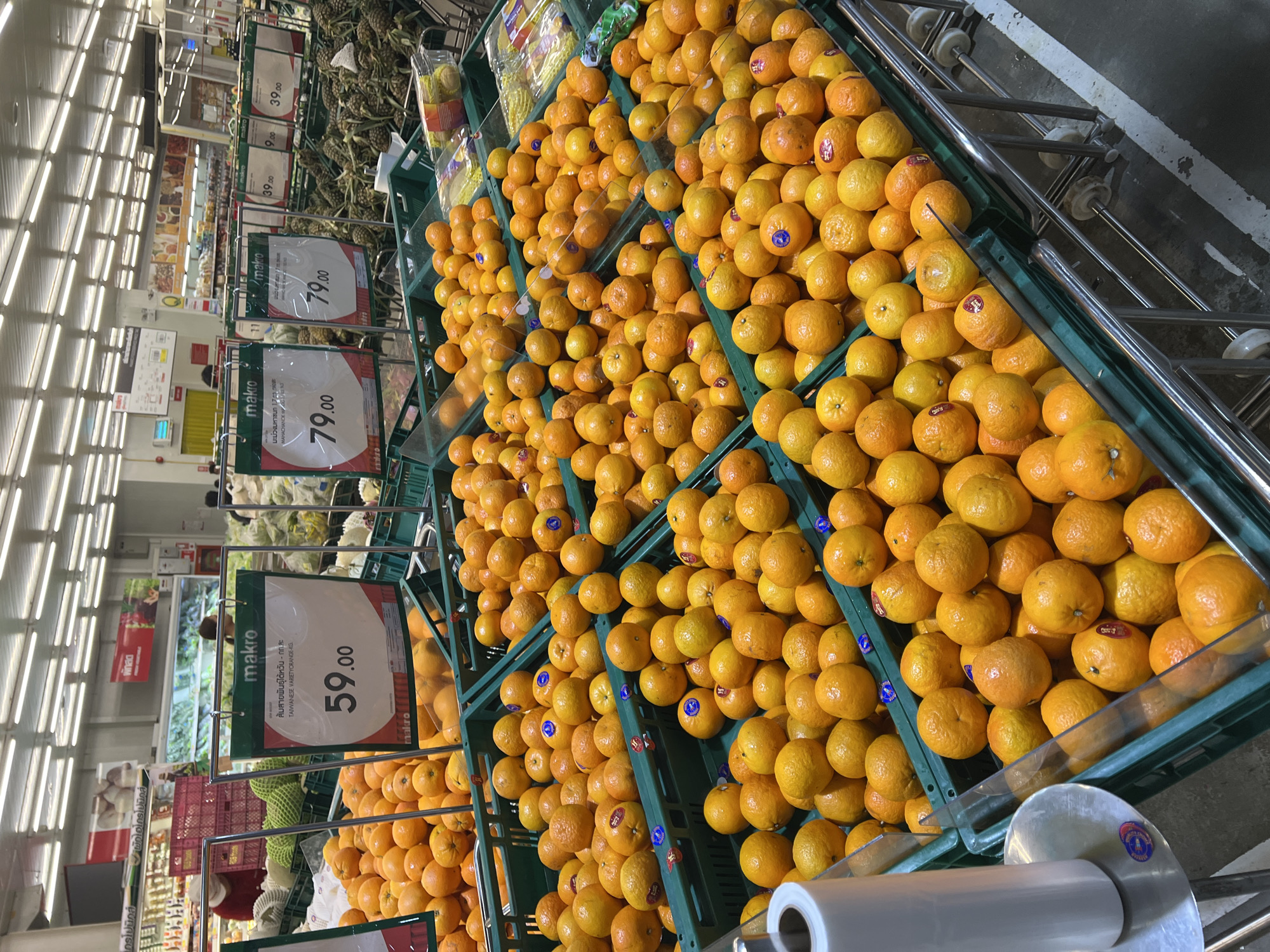 This is a medium shot of a grocery store produce section, specifically showing a display of oranges. In the foreground, green plastic crates are filled with oranges. Some of the oranges have stickers with blue circular logos. To the left, a roll of clear plastic wrap on a metal stand is partially visible. Behind the oranges, a white sign with "makro" logo is hanging, and also price tags with "59.00" and "79.00" written on them. In the background, other produce such as durian and pineapples are visible, as well as shoppers browsing the aisles. The lighting appears to be bright, typical of a supermarket. The store is located in Baan Bo Phut, Thailand.