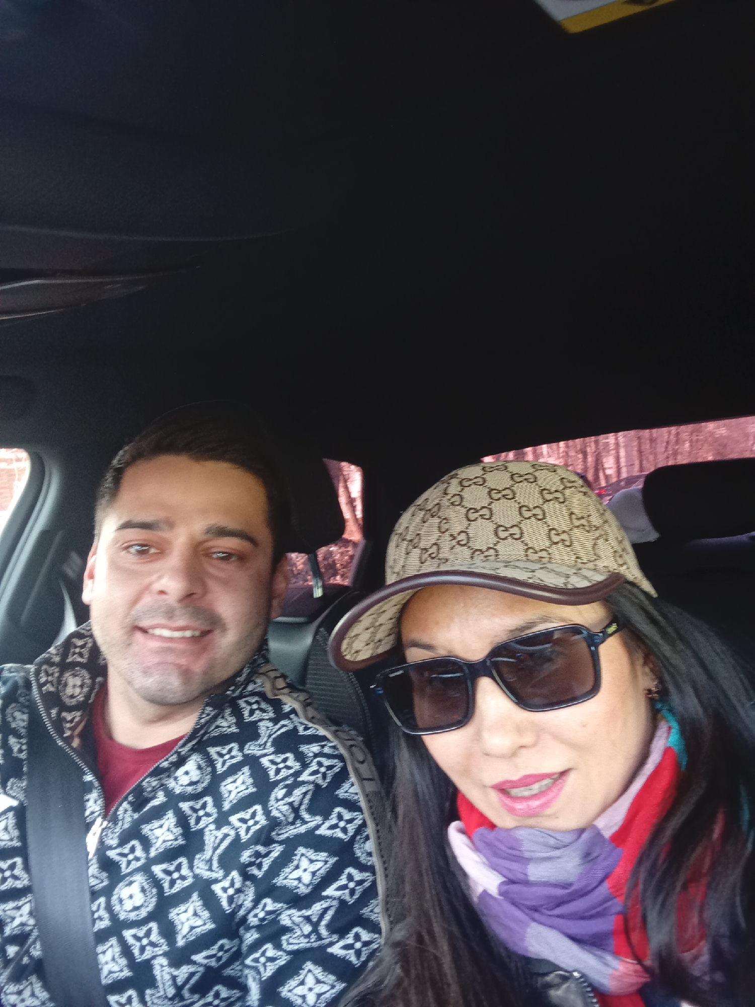 This is a daytime selfie taken inside a car, featuring two individuals: a man and a woman.
The man, positioned on the left, smiles broadly at the camera. He has short dark hair and is wearing a dark jacket with a repeating white pattern resembling the Louis Vuitton monogram. A red garment is visible underneath his jacket, and he has a seatbelt fastened across his chest.
The woman, closer to the camera on the right, is wearing a beige baseball cap with a distinctive repeating "GG" pattern, characteristic of the Gucci brand. Her eyes are obscured by dark sunglasses, and she has long, dark hair. A multi-colored scarf, featuring shades of red, purple, and blue, is wrapped around her neck. Her lips are painted with bright pink lipstick, and she exhibits a relaxed, smiling expression, posing for the camera.
The car's dark interior, including the headliner, is visible. Through the windows in the background, a blurred outdoor scene of trees can be seen, suggesting a natural environment, possibly in a suburban or rural area like Izhevsk, Russia. The bare branches of the trees imply a cooler season, such as late autumn or winter. The overall mood is casual and happy.
