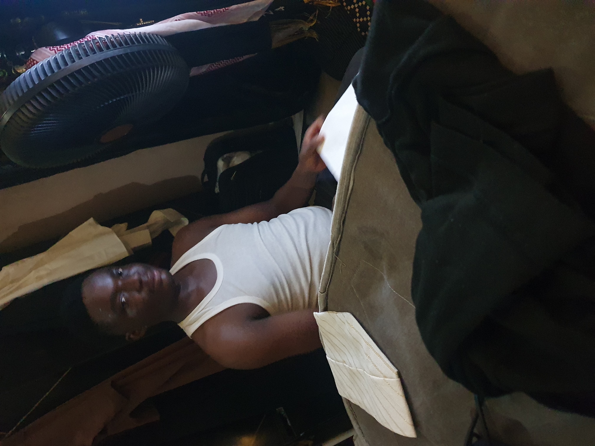 A man is lying on a dark surface, possibly a floor or couch, in a dimly lit indoor environment. He is wearing a white tank top. Beside him is a light-colored object or piece of fabric with thin stripes, potentially part of a garment. A fan is visible in the upper-left corner of the image. The location is unspecified, the city of Lokoja, Nigeria isn't confirmed.