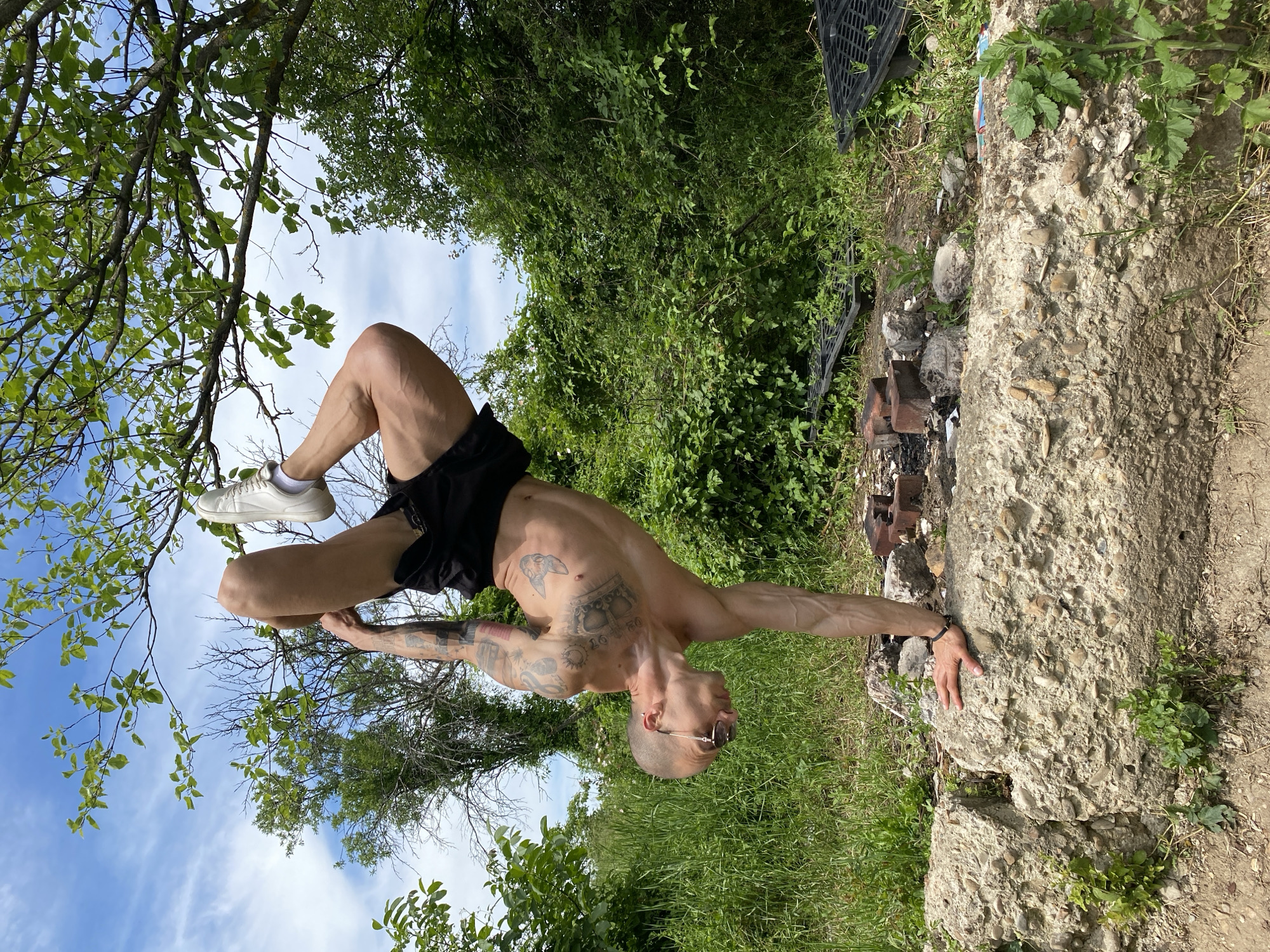 A shirtless man wearing black shorts, white sneakers, and glasses performs a yoga pose outdoors, supported by a tree branch and a concrete structure. The location appears to be a park or overgrown area near the city of Pleven, Bulgaria, based on the foliage and concrete remnants present in the scene. The man has tattoos on his torso and arms. The background includes trees, grass, and a blue sky with clouds.