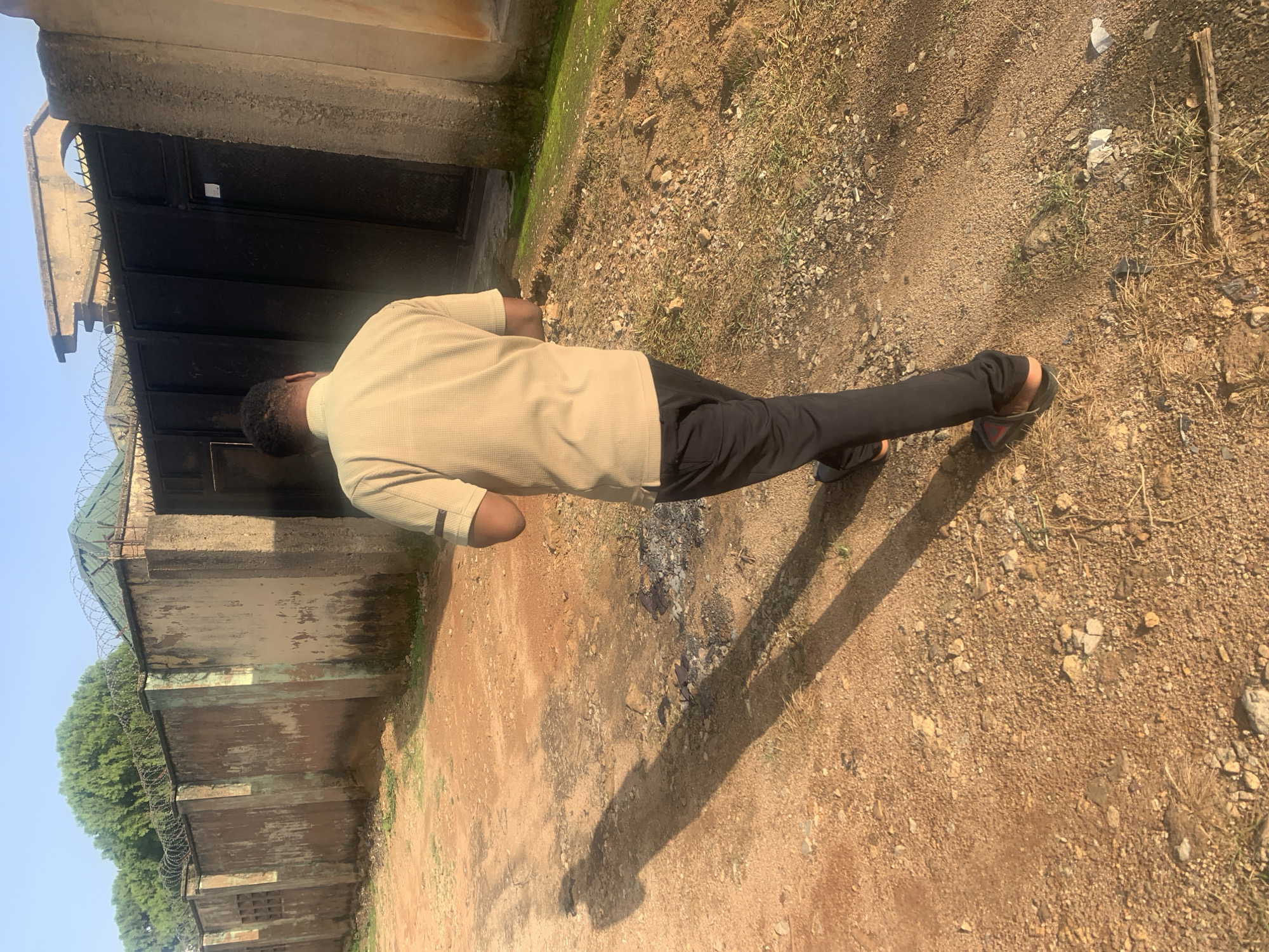 The image depicts a scene on a dirt road in a residential area, possibly in Jos, Nigeria, characterized by an overcast sky and a dry, dusty environment. A man, dressed in a light brown polo shirt and black trousers, walks away from the camera, his posture suggesting he is looking at a mobile device. His shadow stretches long across the reddish-brown, gravelly ground.
The background features a high concrete wall, topped with barbed wire, indicating security measures. Behind the wall, lush green trees are visible. Adjacent to the wall is a large black metal gate, secured with a padlock. Further to the right, a portion of another building, possibly painted in an ochre hue, is visible. The ground is uneven, with scattered stones and patches of dry grass. The overall impression is one of a warm, possibly arid climate, with signs of both residential construction and natural vegetation.