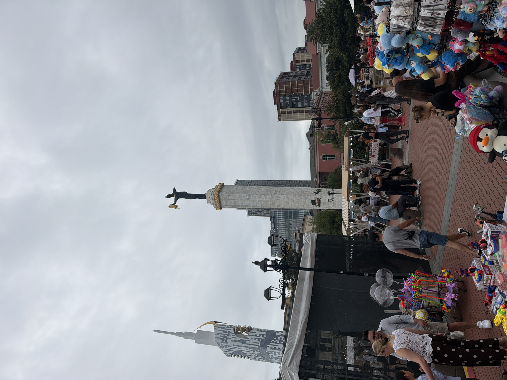 This is an outdoor daytime, medium shot photograph taken in Batumi, Georgia, featuring a bustling public square with market stalls. Dominating the background are architectural landmarks including a tall, modern skyscraper with a distinctive design, and a classical monument topped with a statue holding a golden object. In the mid-ground, several people are seen browsing market stalls that sell various items, including toys and stuffed animals. The ground is paved with red bricks. The sky is overcast. In the foreground, a variety of toys, including action figures and stuffed animals, are displayed for sale on the ground and on tables. People are walking, shopping, and interacting within the square.