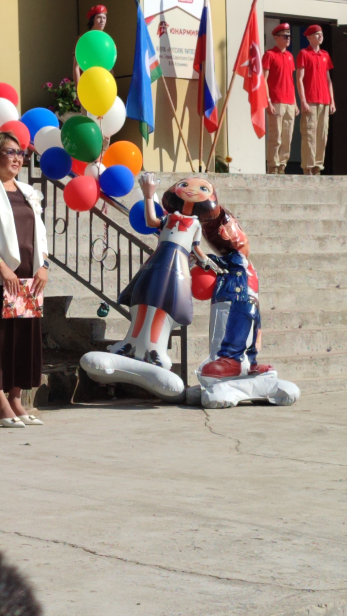 A group of people stand on a concrete outdoor staircase in front of a building in Mirny, Russia. Two young males in red berets and red shirts stand at the top of the stairs. In front of them, two large inflatable figures are present: one of a cartoon character in a blue dress and white sailor collar, and the other of a figure in blue overalls and a red shirt. A woman in a white jacket and dark dress stands on the lower part of the stairs to the left. Several colorful balloons are visible around the inflatable figures and to the left of the woman. Flags are also visible in the background.