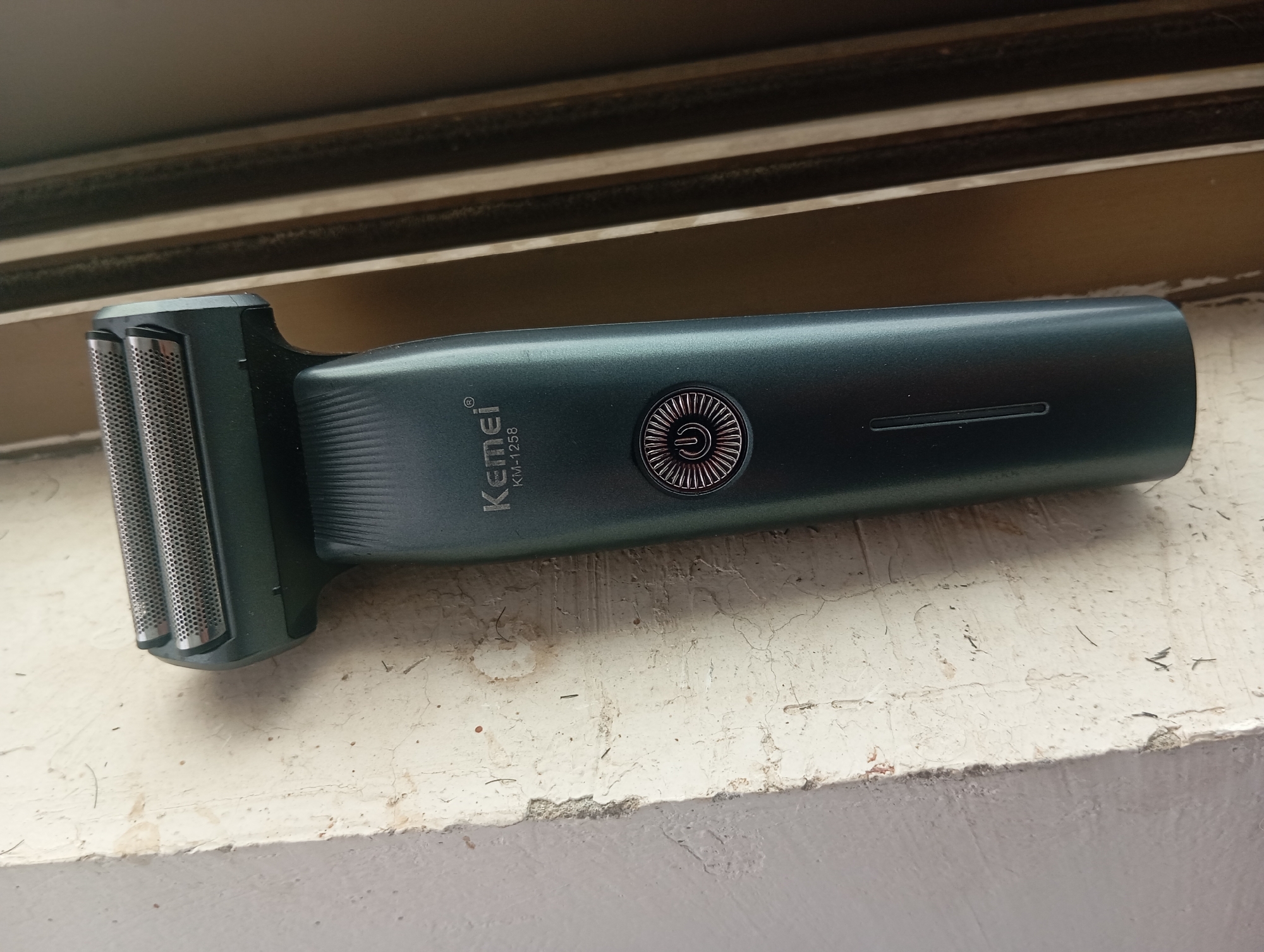 A dark teal electric shaver is placed on a weathered, off-white ledge. The shaver has a brushed metal foil head with two rectangular cutting sections. The body of the shaver is smooth with a subtle ribbed texture near the head. A circular power button with a stylized power symbol is located on the side of the body. Text printed on the shaver reads "Kemei" and "KM-1258". Above the ledge, there are horizontal wooden slats, possibly part of a window frame. The surface of the ledge shows cracks and is speckled with dirt and some dark hair-like fibers. The image is taken from a slightly elevated, eye-level perspective.