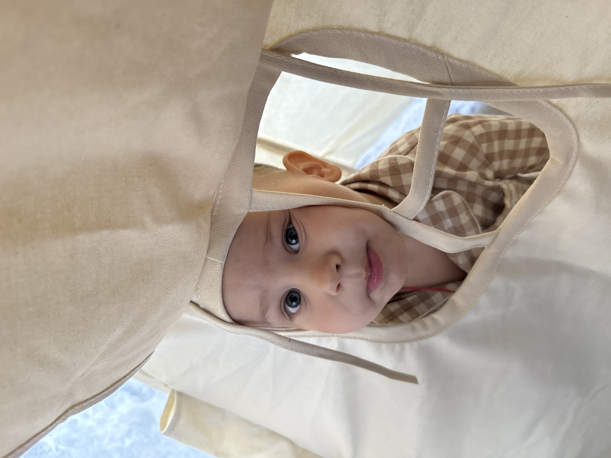 A baby is looking downward through a beige-colored cloth object that appears to be a container or tent. The baby has fair skin, blue eyes, and is wearing a gingham-patterned garment in shades of brown and white. The cloth object has a drawstring visible. The background appears to be a light sky. The city of Заволжье, Russia is not determinable from the image.