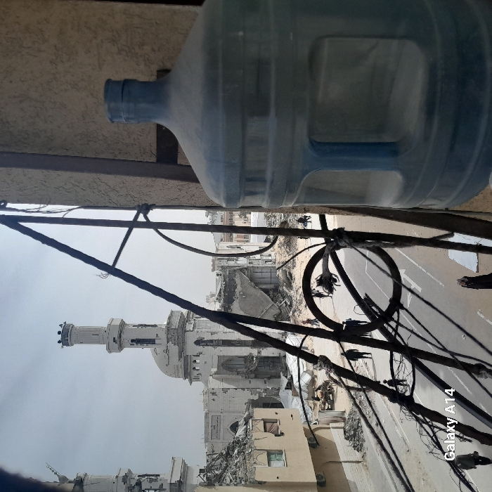 The image depicts a street in Khan Yunis, Palestinian Territory, from an elevated perspective, showcasing widespread urban destruction under an overcast sky. In the foreground on the right, a large, dusty, translucent blue plastic water cooler bottle is partially visible. Dark, rusty metal bars and wires, possibly from a balcony or utility infrastructure, traverse the mid-ground, segmenting the view.
The central background features a severely damaged large white mosque, with one prominent minaret standing largely intact despite visible wear. A second minaret is partially visible to the left, also appearing damaged. The main structure of the mosque, including its dome and roof, is heavily compromised, with extensive sections reduced to rubble and internal architectural elements exposed. Surrounding the mosque are multiple multi-story buildings, many of which are partially or entirely collapsed, resulting in large accumulations of debris.
A paved street with white road markings runs horizontally across the mid-ground, flanked by areas covered in rubble and dirt. Several individuals are present on the street and amidst the debris, engaged in walking or standing. A small group of people is discernible near the damaged mosque, close to piles of rubble and white tarps laid on the ground. The overall environment is characterized by extensive structural damage and debris. A "Galaxy A14" watermark is present in the lower-left corner.