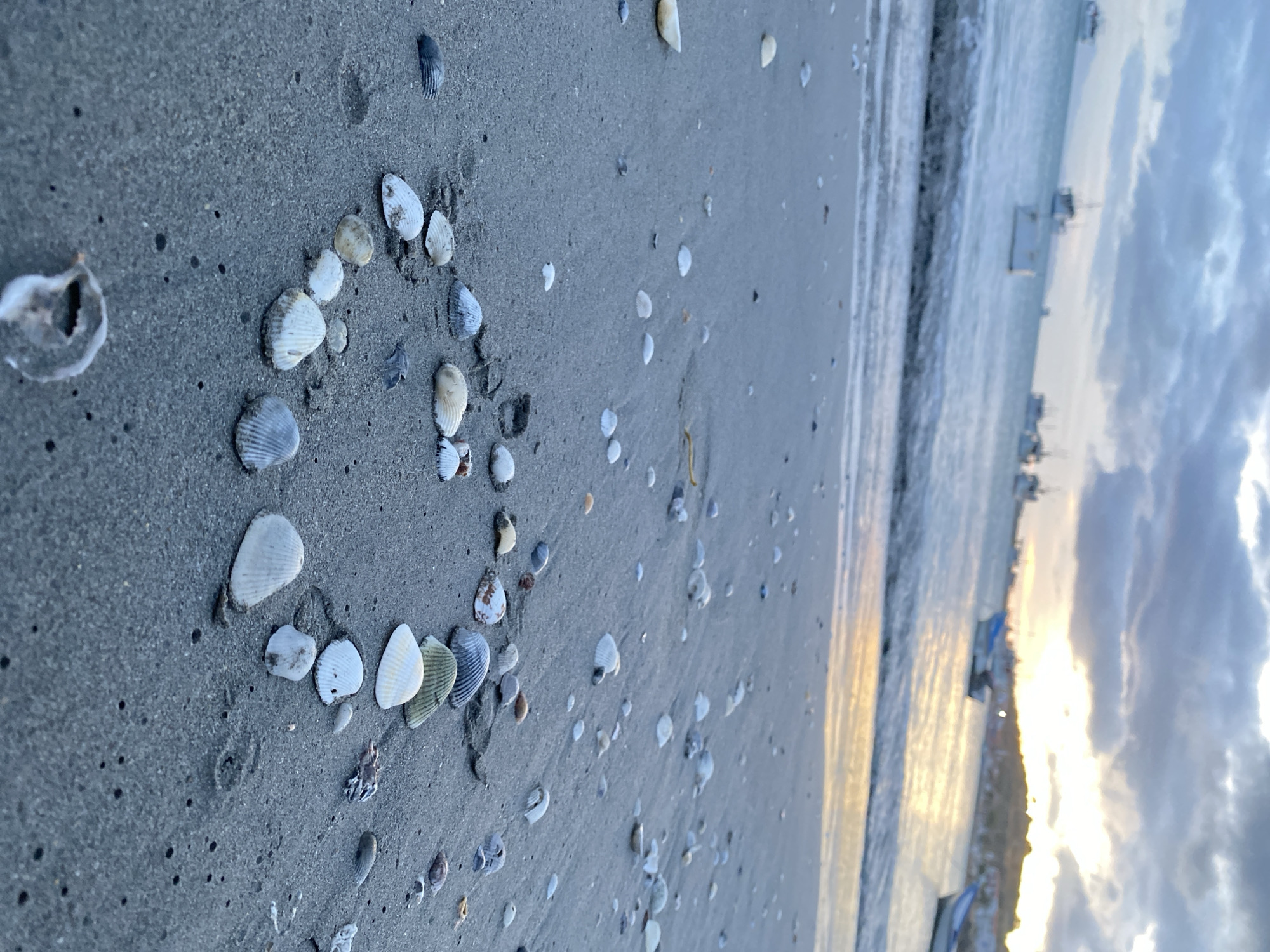 The image captures a beach scene in Juan Griego, Venezuela, likely taken near sunset based on the lighting. A heart shape is formed on the sand using various seashells. Further from the heart are scattered shells. The shoreline shows gentle waves. In the distance, boats are visible along the coast. Above, there's a cloudy sky with colors indicating the approaching sunset.