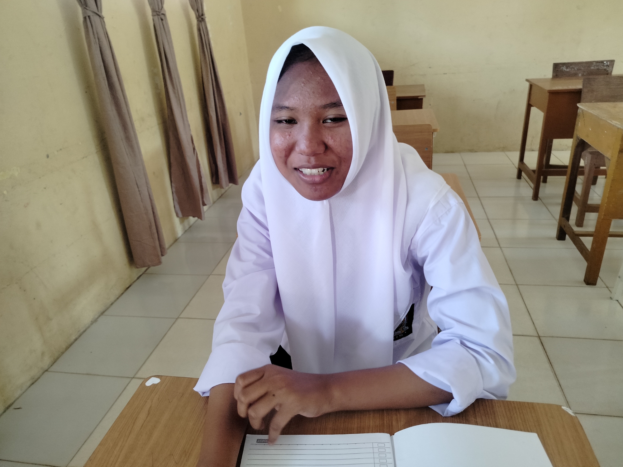 A young person wearing a white hijab and uniform sits at a wooden desk in what appears to be a classroom in Purwodadi, Indonesia. The individual is looking directly at the camera with a slight smile and is positioned in the foreground. Behind them, several other wooden desks are visible, along with a wall with hanging drapes and tiled flooring. The overall impression is of an everyday school scene.