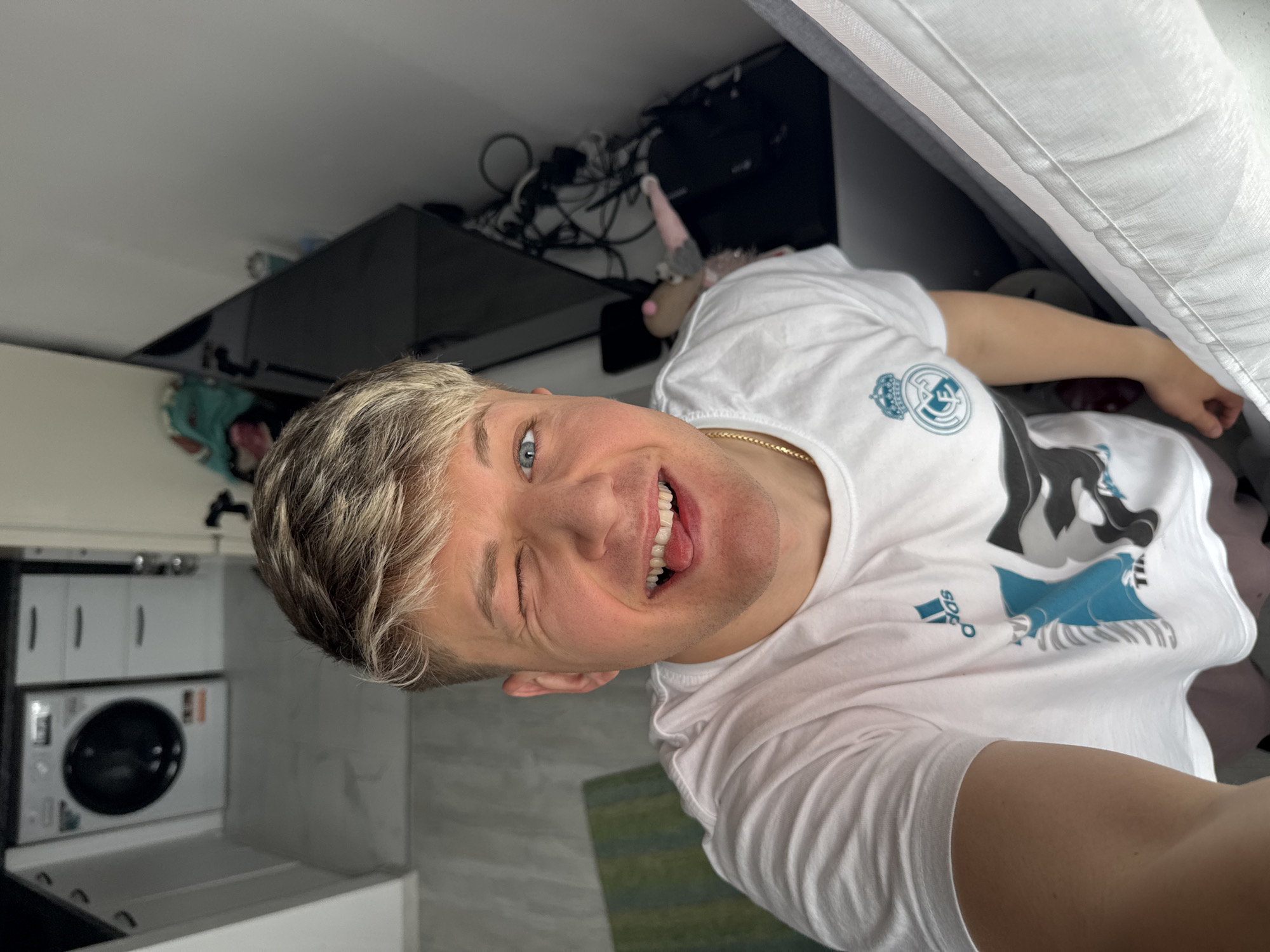 A selfie image shows a fair-skinned individual with blond hair and blue eyes, winking at the camera and sticking out their tongue. They are wearing a white t-shirt with a Real Madrid logo and an Adidas logo. A thin gold chain is visible around their neck.
The background appears to be the interior of a home or apartment. In the top left corner, there are white kitchen cabinets with a washing machine. Behind the person and slightly above them is a cluttered area with a television screen and tangled cables. Part of a white upholstered bed or sofa is visible in the upper right corner. The floor appears to be gray and there's a green rug visible.
There is insufficient information in the image to establish the location in Plovdiv, Bulgaria.