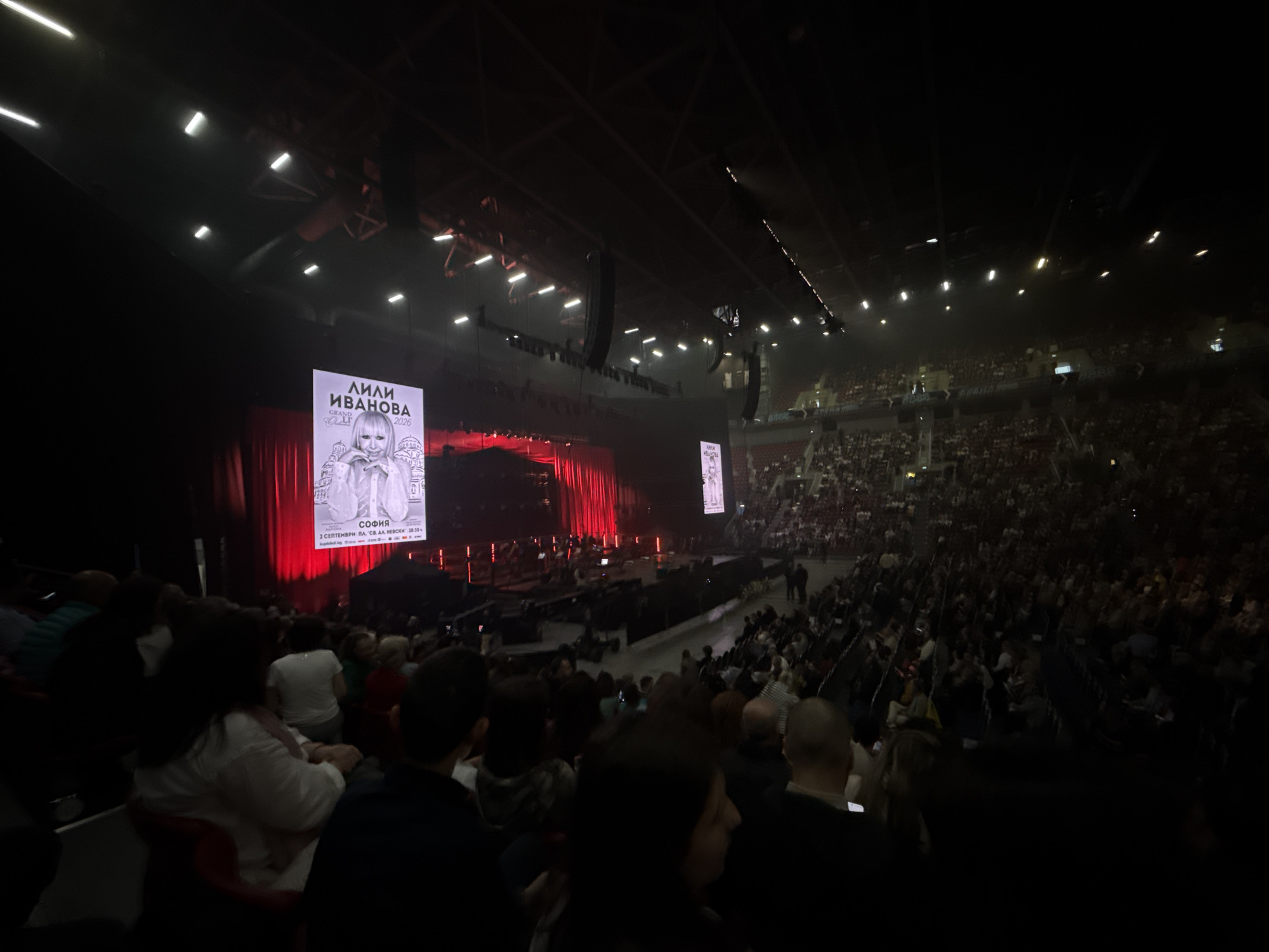 The image shows a concert of Lili Ivanova in Sofia, Bulgaria. The concert is held on September 2nd on "St. Alexander Nevsky" square, based on the banner, advertising the event. The view is from the audience, looking towards the stage with red curtains. The stage has the banner of Lili Ivanova's tour 2026, and is lit up. An orchestra can be seen on the stage. The venue is crowded with people watching the concert. The room is dark, lit up by the stage and venue lights.